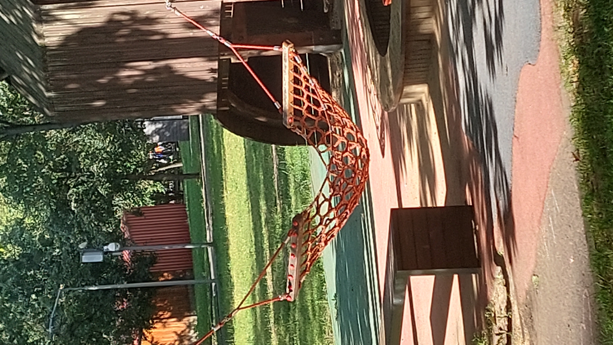 A playground scene is depicted in Moscow, Russia, on a sunny day. In the foreground, a dark brown wooden bench is positioned on a red-colored surface. Further back, a hammock made of orange netting is suspended by red ropes from a dark wooden structure, possibly part of a playhouse or climbing frame. The hammock appears to be empty. In the background, green grass, trees, and a tall lamp post with security cameras are visible. A red-colored fence is also present. The overall setting suggests a public outdoor recreational area.