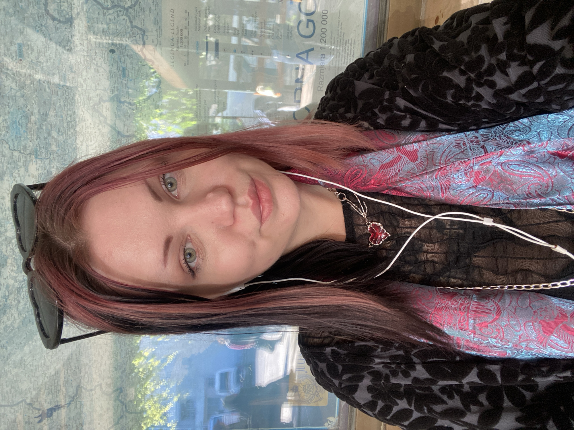 A portrait of a woman with long, pink-tinged hair and fair skin is captured indoors with a window as the backdrop. She is wearing a black top with a decorative pattern and a colorful scarf with a paisley design. A pair of sunglasses is partially visible on her left. She has earbuds in. A necklace with a heart-shaped pendant is seen. The window reveals an outdoor scene, and a map with the heading "LEGENDA-LEGEND" is affixed to the window. Text reading "Razmjera 1:200 000" is also visible. The map indicates the location as Montenegro, specifically in Budva.