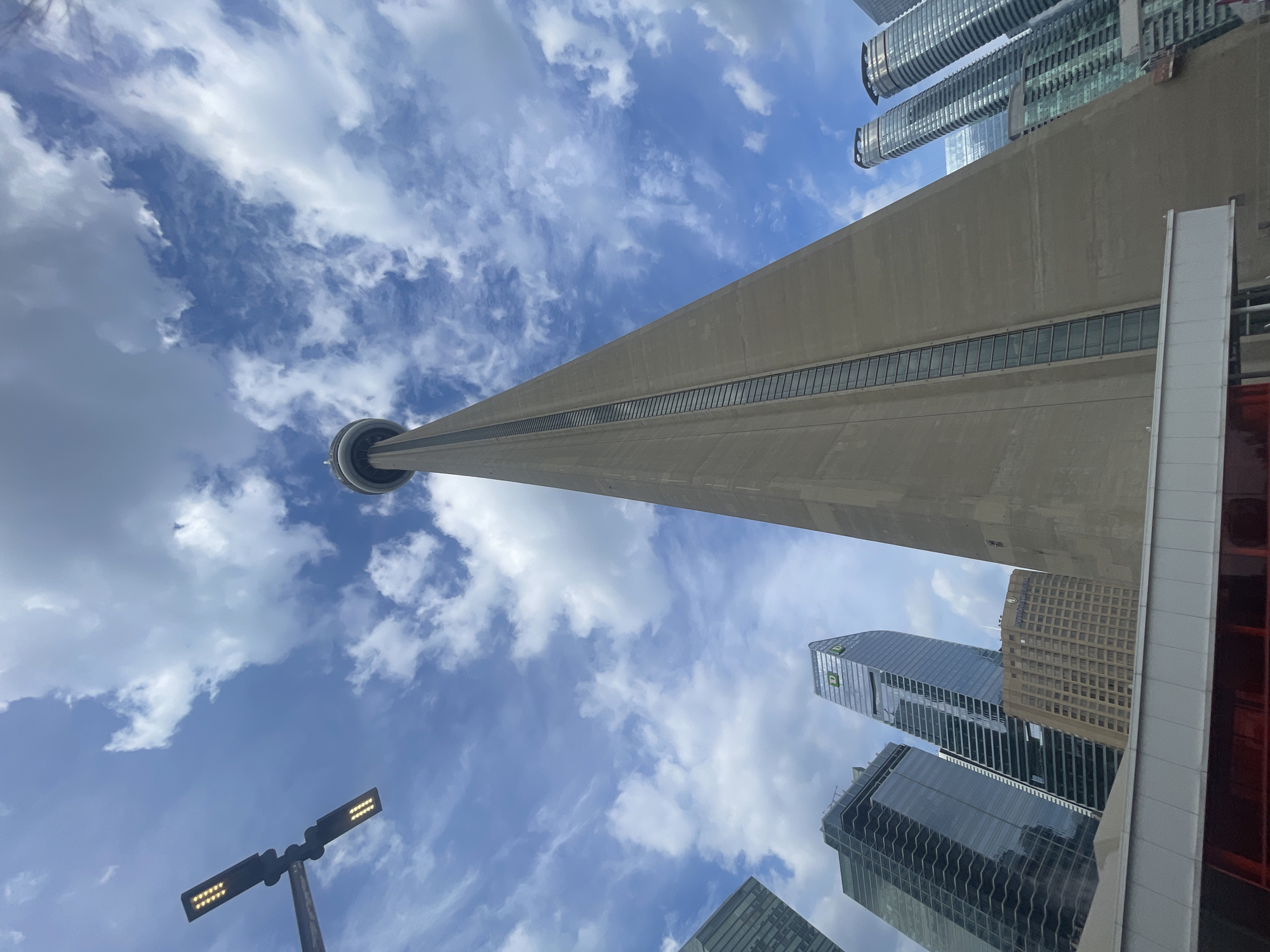 The image shows a low-angle view of the CN Tower in Toronto, Canada, against a partially cloudy sky. The concrete tower dominates the center of the frame, tapering as it ascends. To the left of the CN Tower, several modern skyscrapers are visible, characterized by glass facades that reflect the sky. One of these buildings features the prominent green TD logo. In the foreground, a white elevated walkway with a red structure underneath is partially visible. A street lamp with multiple LED lights is positioned in the upper left corner of the image. The environment appears to be an urban setting with clear daylight.