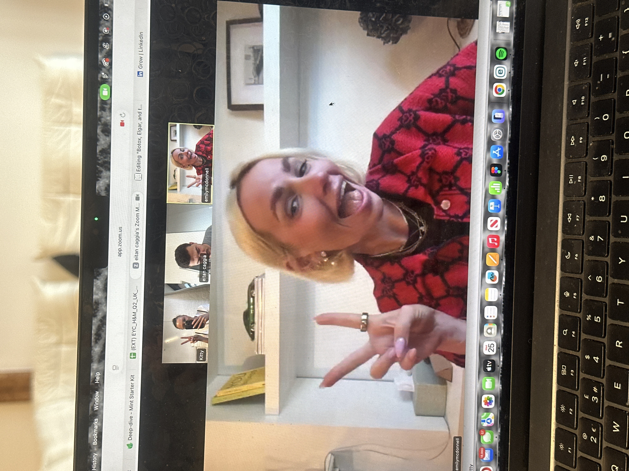 The image displays a close-up of a black Apple MacBook laptop screen, partially obscured by the laptop's keyboard in the foreground. The screen presents an active Zoom video conference featuring four participants. The prominent participant, labeled "emilymcdonnell," is a blonde woman with short hair, wearing a red and black patterned jacket, multiple gold necklaces, and pearl earrings. She is smiling broadly with an open mouth and forming a peace sign with her left hand, which displays light pink manicured nails and a gold ring on the ring finger. Her background consists of a white shelving unit holding a yellow book, a green model car, and a framed picture. In the smaller top frames, "Kitty" is visible, holding a smartphone and also making a peace sign, while "eitan caggia" appears to be looking downwards.
The macOS dock at the bottom of the screen shows numerous application icons. Several Safari browser tabs are open at the top, one of which is titled "(EXT) EYC_H&M_Q2_UK_...". The laptop's keyboard features a QWERTY layout with the "£" symbol present on the '3' key. The menu bar displays "Wed 25" and icons indicating Wi-Fi and battery status, alongside notification counts for Mail (18,860) and Messages (57). The immediate environment behind the laptop is a blurry background of light-colored fabric and a wooden element. This scene is located in the City of London, United Kingdom, evidenced by the "£" symbol on the keyboard and the "UK" reference in a browser tab.