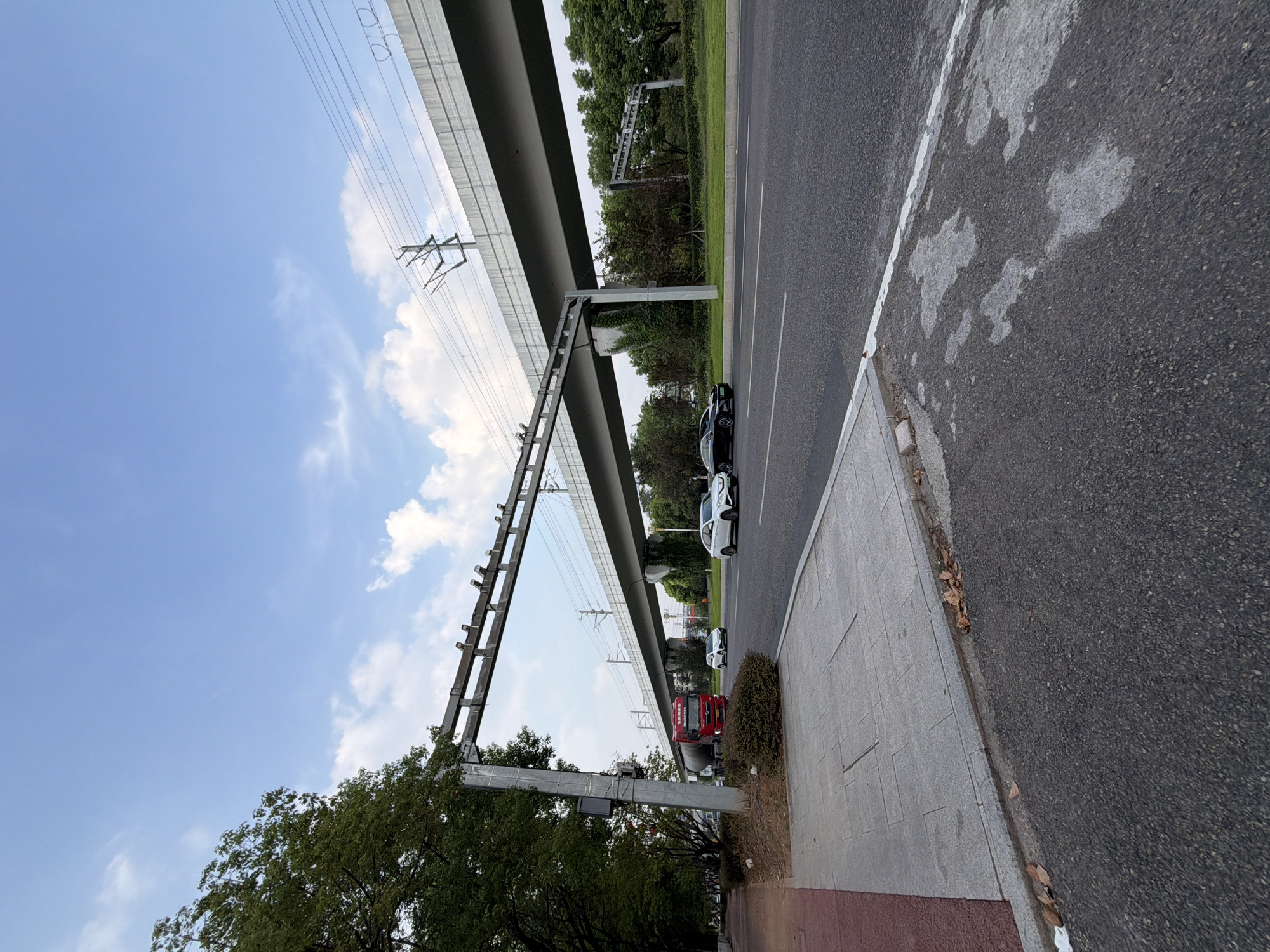 An elevated train track runs overhead, supported by concrete pillars, with power lines crisscrossing the sky. Below the tracks, a two-lane road stretches into the distance, bordered by a sidewalk on the left and trees and greenery on the right. A red truck is parked on the side of the road, and several cars are driving on the road. The sky is blue with scattered white clouds. The scene appears to be in the city of Jiangjin, China.