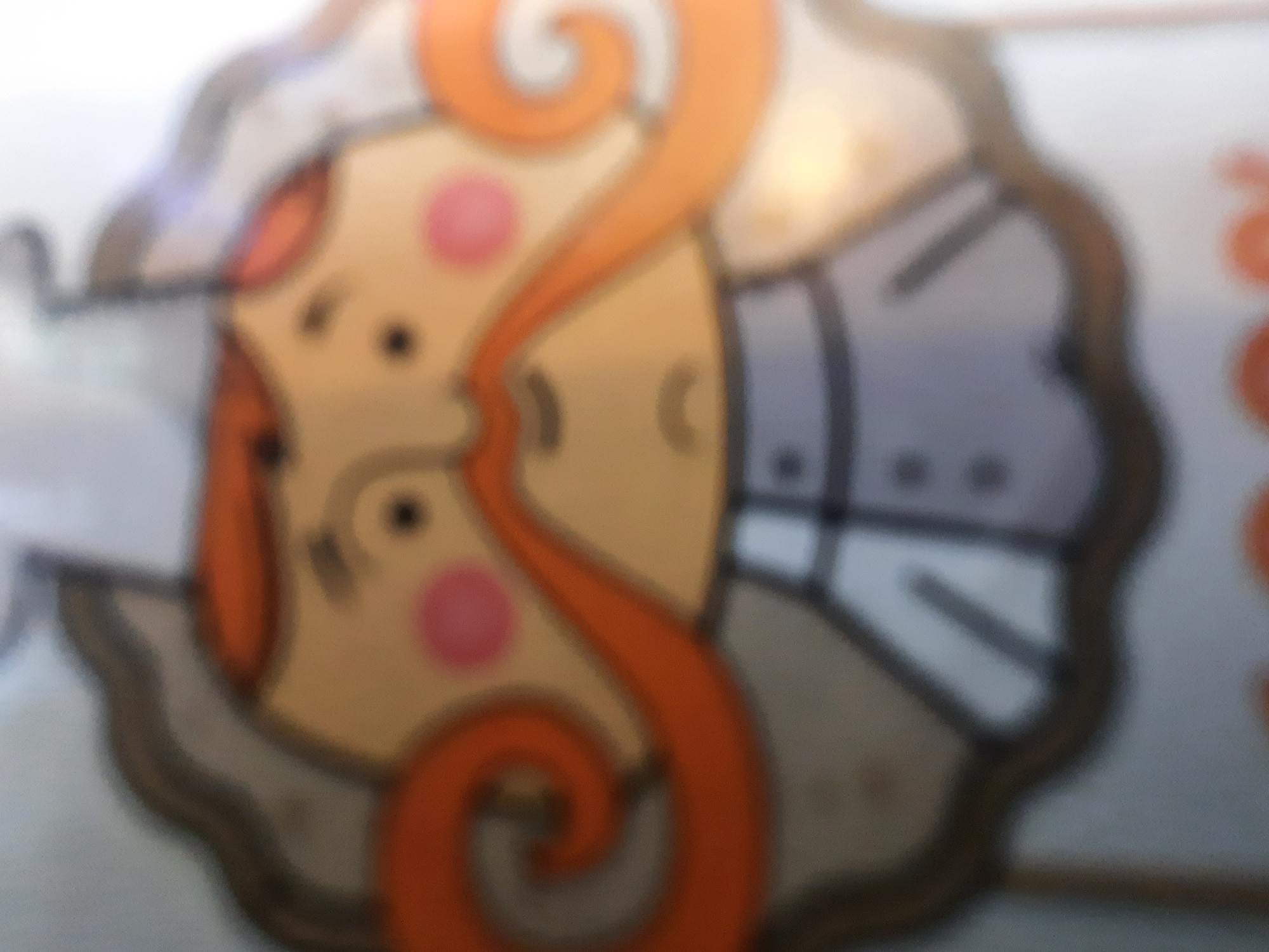 A blurry close-up shows a cartoon depiction of a red-haired person with rosy cheeks, wearing what appears to be a uniform or formal attire. The image is on a surface, possibly a container or packaging. The city and location are unknown due to the blurry nature of the image.