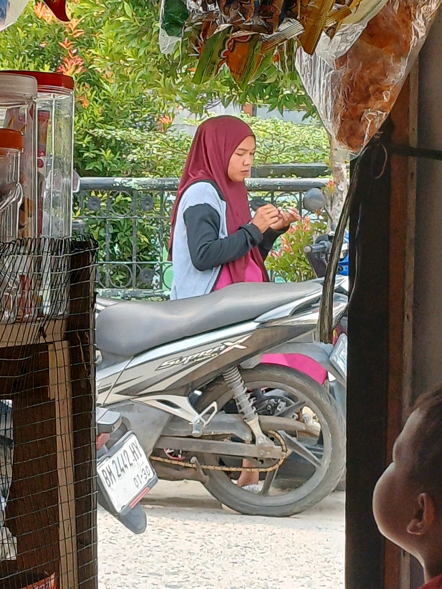 A young woman wearing a maroon hijab and a light blue and black long-sleeved shirt is sitting on a motorcycle, looking down at her hands as she holds something. She is in an outdoor setting, with lush green foliage and trees in the background. A black metal fence is visible behind her. To her left, there are large clear jars filled with various items, and a wire rack. The motorcycle's license plate, visible on the rear, reads "BH 2442 HY 01-20". Part of a structure, possibly a stall or shop, is visible on the right side of the image, with wooden beams and a hanging plastic bag filled with brown food items. A child's head is visible in the lower right corner of the frame, facing away from the camera. The ground appears to be a gravel or coarse sand surface. The scene suggests a commercial area, possibly a roadside stall or market in Dumai City, Indonesia, given the license plate format common in that region.