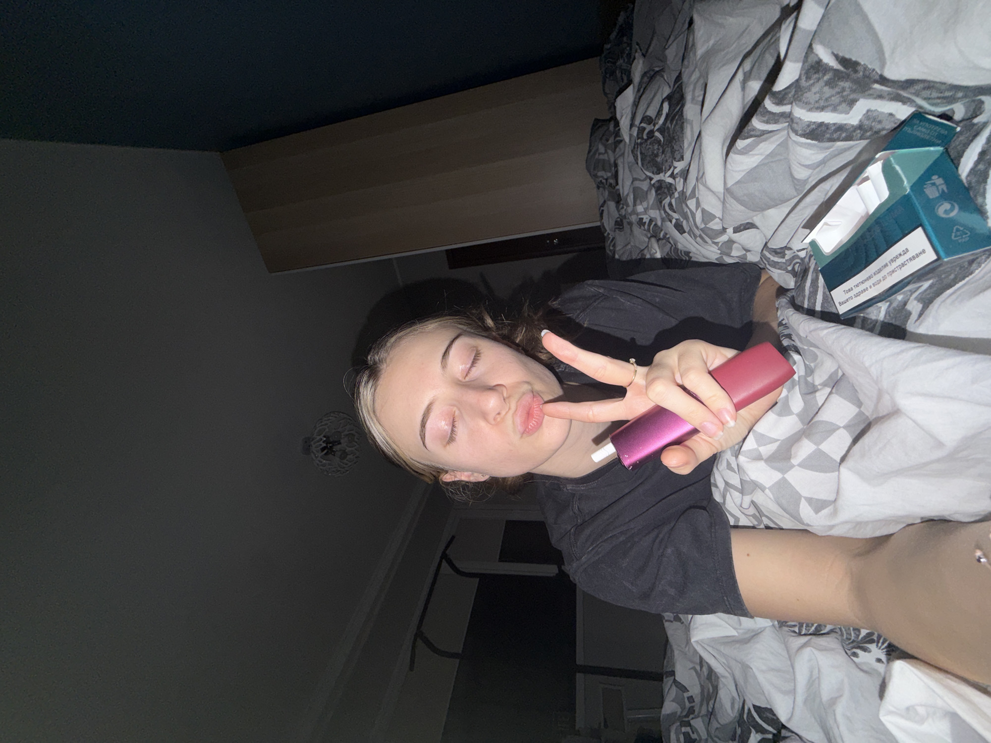 A young woman is lying in bed. She is holding a pink electronic cigarette to her lips and making a peace sign with her other hand. Next to her on the bed is an opened pack of cigarettes. The room is dimly lit. The location is impossible to determine from this image, it can't be identified as being in Plovdiv, Bulgaria.