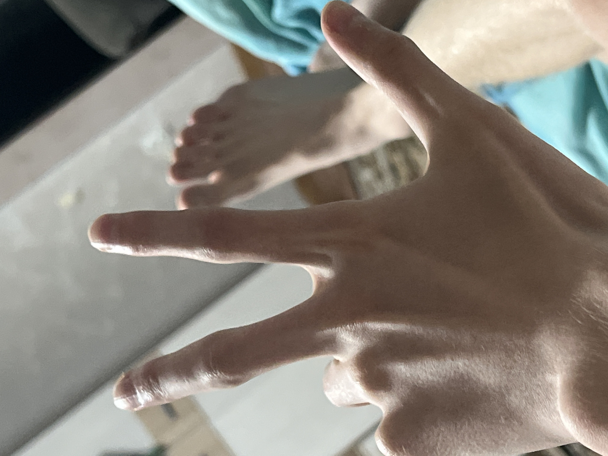 A close-up, low-angle shot focuses on the back of a person's hand and forearm, which is positioned in the foreground. Three fingers are extended upwards: the index, middle, and ring fingers. The index and middle fingers are held together, while the ring finger is slightly separated. The skin on the hand appears fair and slightly oily, with visible veins and knuckles. In the background, out of focus, are a person's bare feet, suggesting they are lying down. A blue cloth is visible in the lower right corner. The environment appears to be indoors, with a neutral-colored wall or ceiling in the upper portion of the frame. There is no visible indication of the city of Gorshki, Russia, within the image.