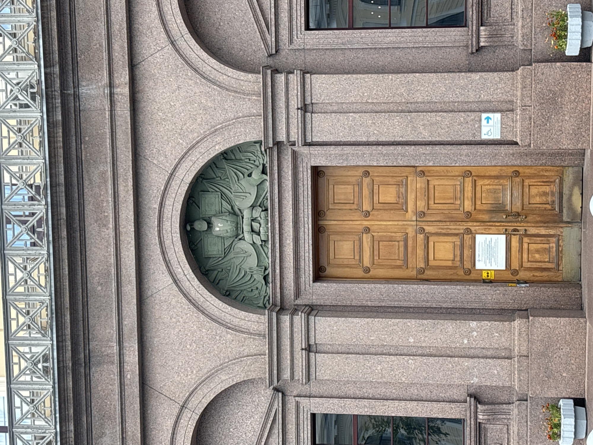 The image shows the main entrance to the Russian National Library in Saint Petersburg, Russia. The entrance features a large, double wooden door with a classical pediment above it. The pediment contains a bas-relief sculpture depicting a figure seated with drapery and symbols of learning. The building's facade is made of granite, and two arched windows are visible on either side of the entrance. A metal railing runs along the top of the building. In front of the entrance, there are two planters with flowers. A sign indicating accessibility for people with disabilities is posted on the right side of the door.