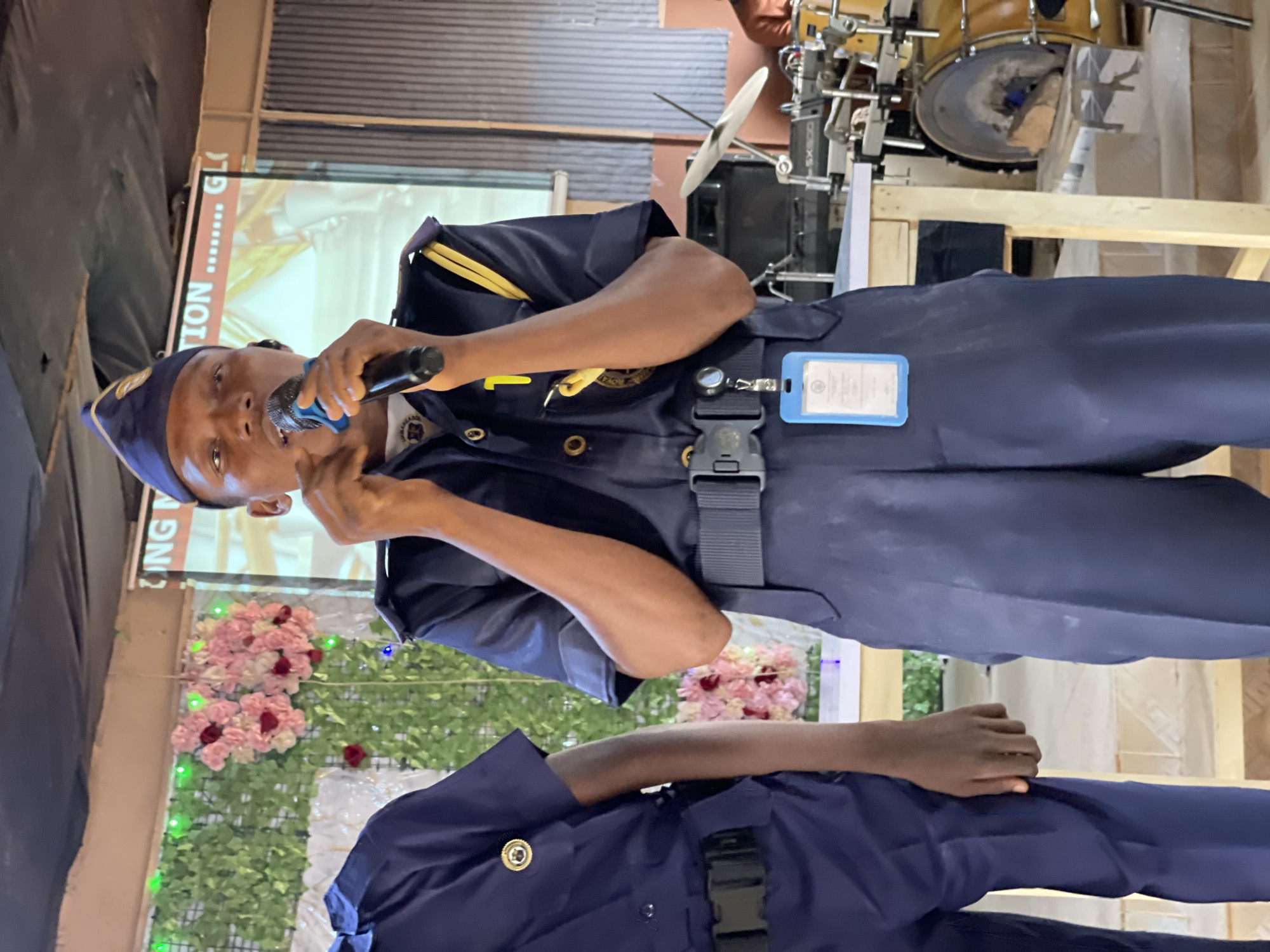 The image shows a man in a dark blue uniform speaking into a microphone in what appears to be an indoor setting. The uniform includes a cap, epaulettes with yellow stripes, a buckle, and an identification card holder. The man is of African descent.
In the background, there's a projection screen displaying text and images, and a flower arrangement adorns the base of what seems to be a makeshift stage. Further back, drum set is placed on a raised platform. Another individual in a similar uniform stands partially visible in the foreground.
Without additional information, confirming the location as Akwanga, Nigeria, is impossible.