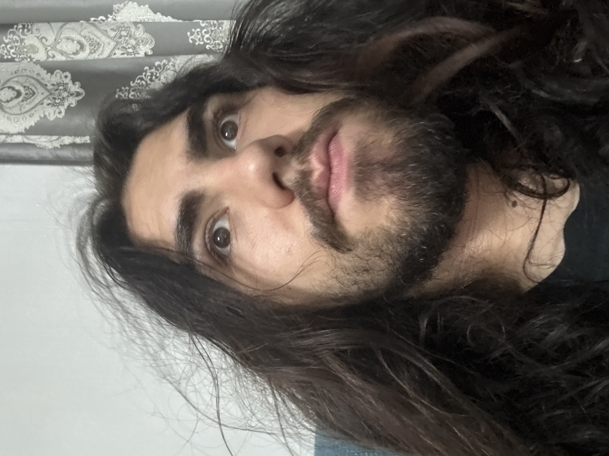 A close-up, eye-level, medium shot captures the face and upper chest of a person with long, dark, wavy hair that is windswept. The subject has brown eyes that are looking upwards and slightly to the left. They have a mustache and beard. The person is wearing a black t-shirt. The background consists of a plain, light-colored wall on the left and a grey curtain with a white, ornamental pattern on the right. The image is well-lit and in focus.