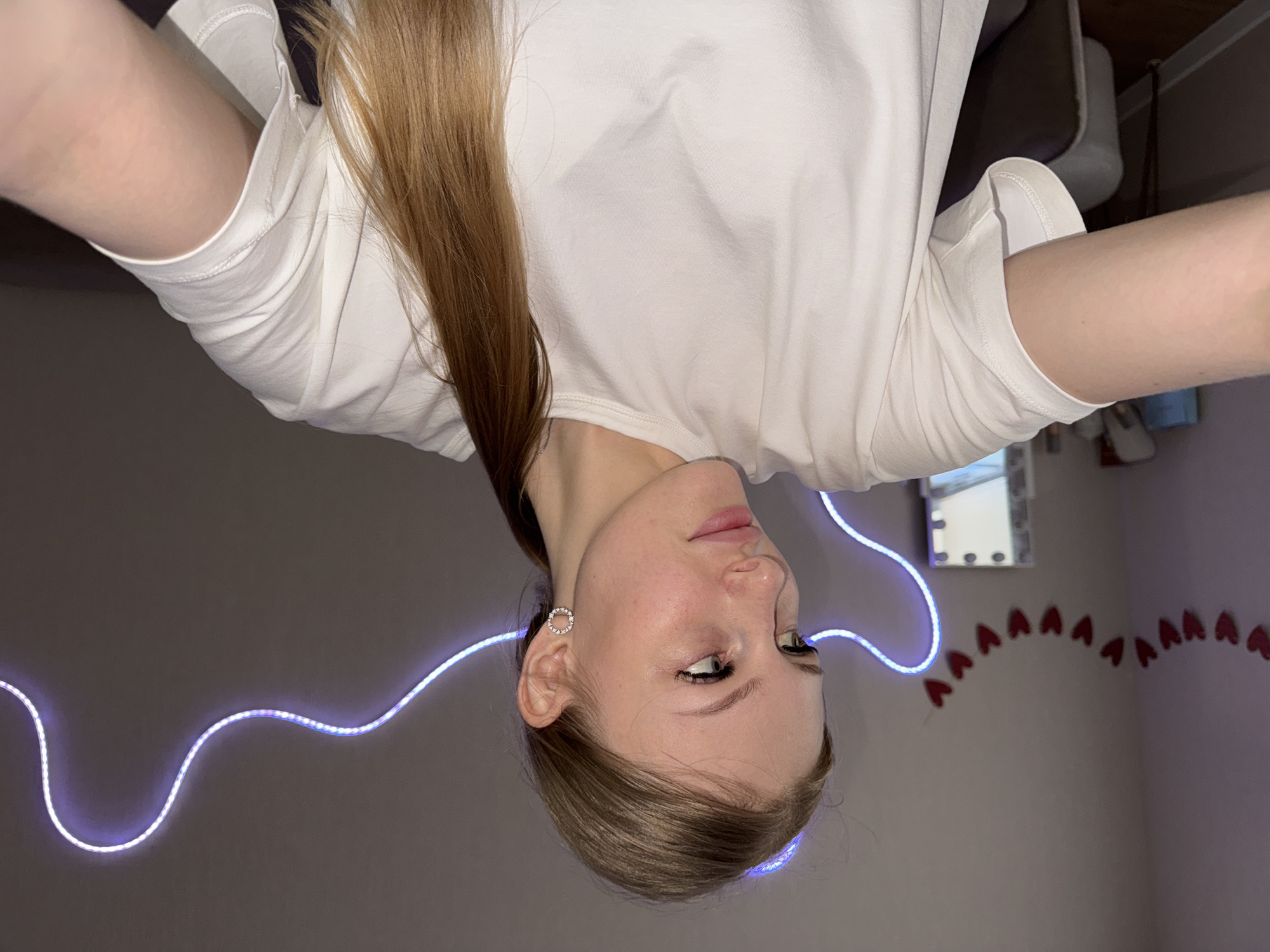 A young woman with long, blonde hair is pictured from a high angle, with her head tilted down. She is wearing a white t-shirt and a circular earring with small stones. A blue LED strip light curves around her and across the wall behind her. On the wall, a string of red heart-shaped decorations is visible. The woman's expression is neutral, and she appears to be looking slightly to her right. The lighting suggests it is nighttime or a dimly lit indoor space. The background includes a blurred reflection of what appears to be a mirror on the wall. The scene is set in a room with grey walls. No visible text is present in the image.