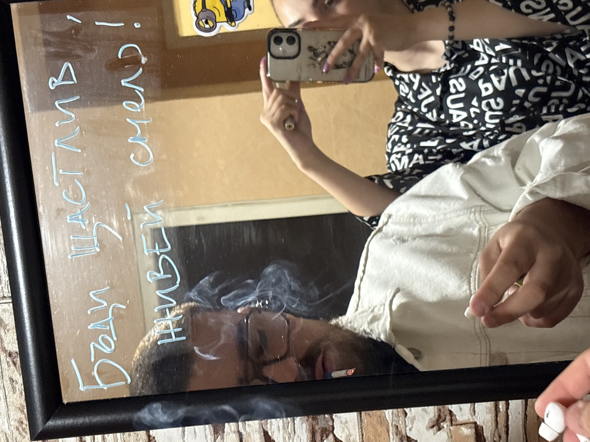 A selfie is taken in a mirror, reflecting two individuals and writing on the mirror itself. In the foreground, a young man wearing glasses and a light-colored jacket is visible, with smoke emanating from a cigarette he is holding in his mouth. He appears to be looking down at something in his hands. To the right, a young woman's arm is extended, holding a smartphone in her left hand as she takes the photo. She is wearing a black and white patterned garment. On the mirror, written in blue marker, are the Bulgarian phrases "Бъди щастлив" (Be happy) and "Живей смело!" (Live bravely!). Behind the individuals, a wall with a brick pattern is visible, and a yellow background with a Minions sticker is also reflected in the mirror. The overall setting suggests an indoor environment, possibly a home, and given the Bulgarian text, the location is specified as Burgas, Bulgaria.