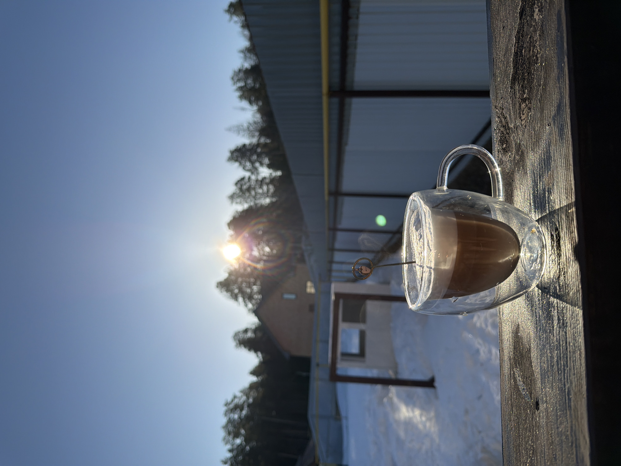 The image shows a glass mug filled with a layered coffee drink, such as a latte or cappuccino. The mug is perched on a dark wooden post, likely part of a deck or fence. Behind the mug, the background reveals a wintry scene with snow-covered ground and a building, possibly a house, partially obscured by trees. A metal structure with a yellow pipe running along it is also visible. The sky is a clear, bright blue with the sun shining and creating a lens flare effect. A thin metal rod with a decorative swirl design is sticking into the drink. The perspective is slightly tilted, suggesting the photo was taken at an angle. Based on the prompt, the location is Chaikovsky, Russia.