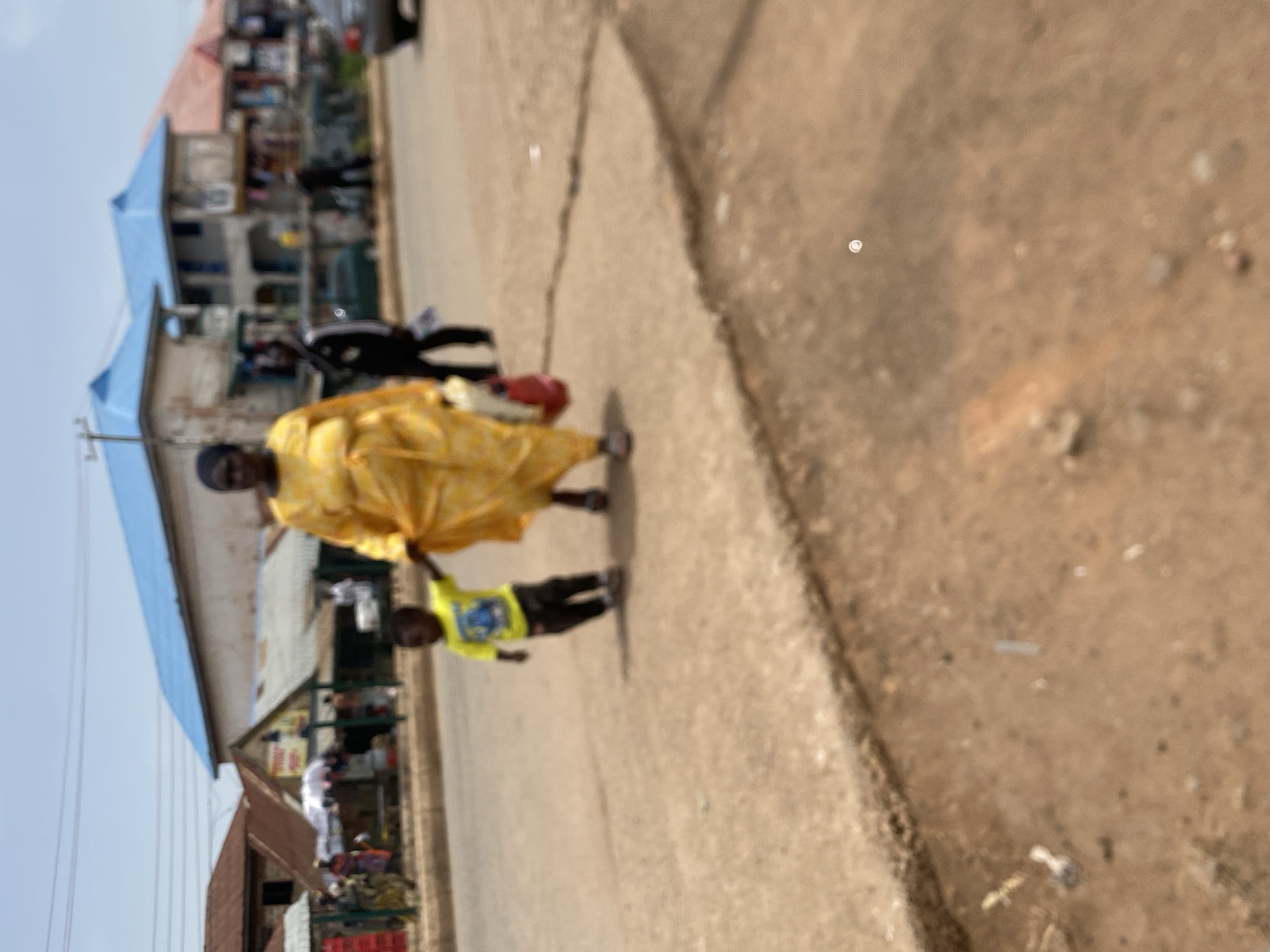 A mother in a yellow garment is walking in the city of Bukuru, Nigeria with a young child wearing a yellow shirt on a city street, possibly on a sidewalk or near a curb. Other structures are present on the other side of the street, including buildings and poles. Pedestrians and vehicles are also in the vicinity. The image is blurry and indistinct.