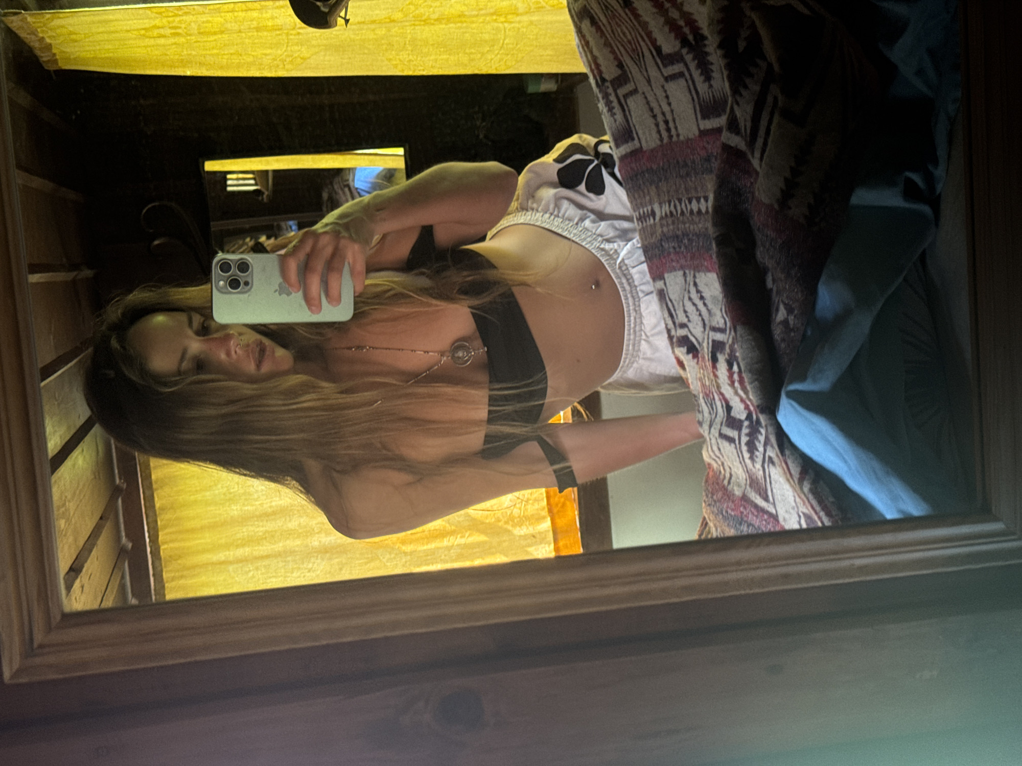 In the city of Unknown, Unknown, a person is taking a selfie in a room. The individual, a female with long blonde hair and a nose ring, is leaning forward towards a mirror. She is wearing a black bandeau top, white shorts with a floral design, and a silver necklace. Her belly button is pierced. The mirror reflects the room, which appears to be dimly lit with yellow curtains visible in the background, suggesting a bedroom or a similar private space. The room's ceiling is made of wooden planks. A patterned blanket is visible in the foreground of the mirror's reflection.
