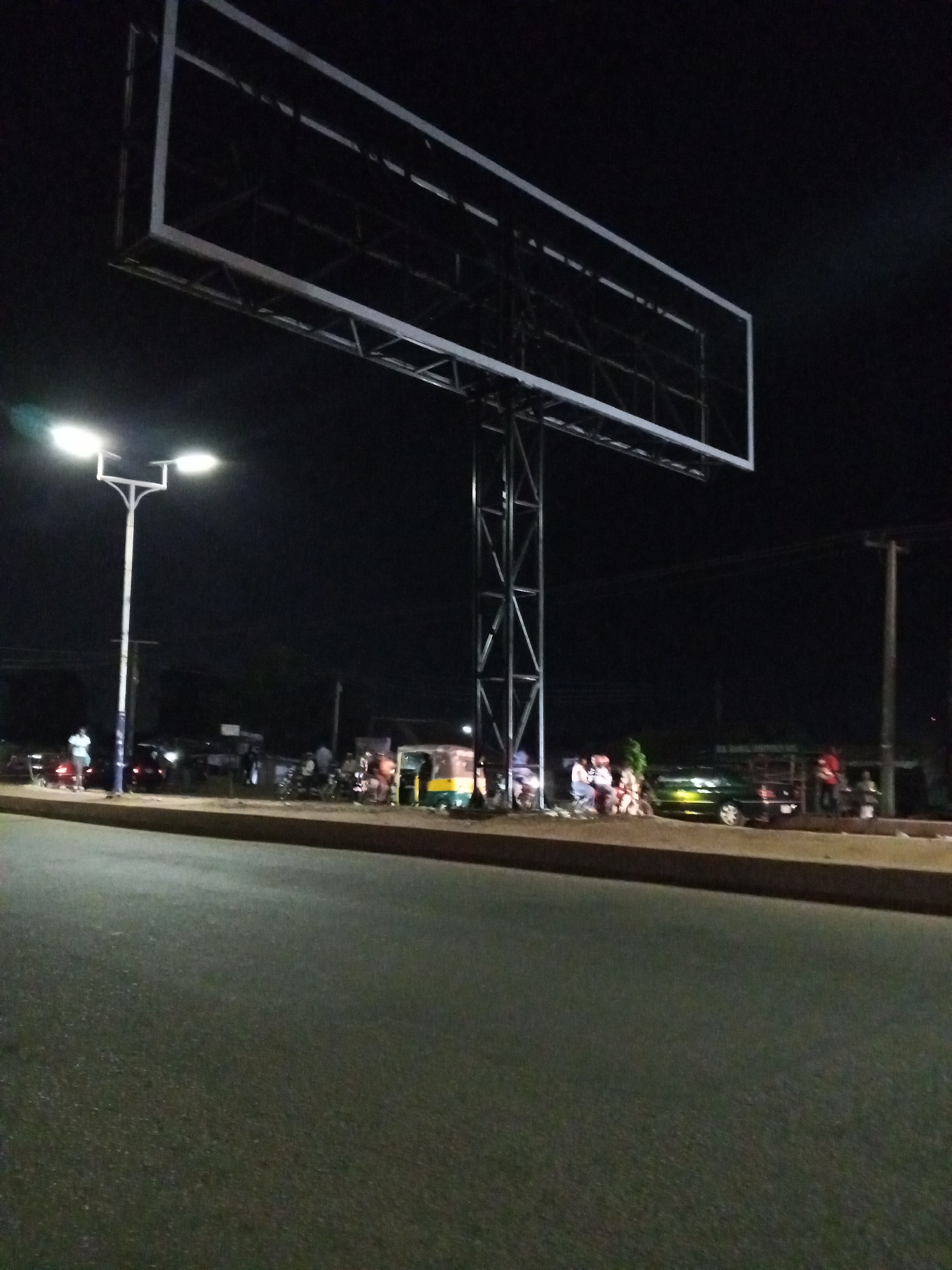 The image shows a street scene in Bukuru, Nigeria, at night. The road surface in the foreground is asphalt, stretching across the bottom half of the frame. A large, empty billboard frame towers over the scene, supported by a tall metal structure. A street light illuminates a section of the scene on the left. Several vehicles and people are visible on the side of the road, including cars, a motorcycle, and a three-wheeled vehicle. People are standing or sitting near the vehicles, suggesting a gathering or waiting area. The background is dark, with obscured buildings and trees.