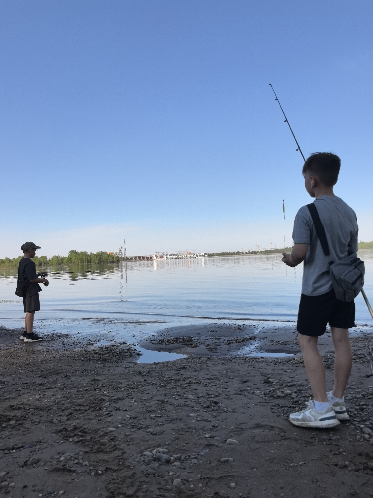 The image depicts two boys fishing on a sandy beach next to a body of water. The sky is clear and blue. One boy, wearing a hat and dark clothing, stands on the left side of the frame, holding a fishing rod. The other boy, seen from the back on the right, wears a grey shirt, black shorts, and white sneakers, also holding a fishing rod. A grey bag is slung over his shoulder. In the background, across the water, is a forested shoreline and a large industrial structure, possibly a hydroelectric dam or power plant. The location is Noviy, Russia.