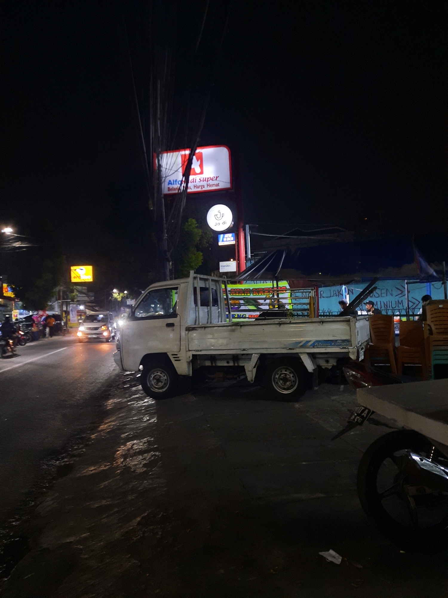 The image depicts a street scene at night in East Jakarta, Indonesia, likely near a commercial area. A white pickup truck is parked on the right side of the road, facing left. The truck bed is empty, and it has "Mega Cargo" written on its side. To the left of the truck, the road is wet and reflects the lights from surrounding signs and vehicles.
Several other vehicles are visible on the road, including a white car with its headlights on, approaching the truck from the distance. Motorcyclists are also present, some parked on the left side of the road.
Above the street, there are numerous signs and electrical wires. One prominent sign, illuminated by red and white lights, reads "Alfa di super Belanja, Harga Hemat," which translates to "Alfa Supermarket, Shopping, Affordable Prices." Another smaller circular sign with the letter "J" inside a circle is also visible. Further down the street, more illuminated signs can be seen, including one that appears to be a bank ATM sign.
The background shows the facades of buildings and awnings, suggesting a busy commercial strip. The overall atmosphere is that of a bustling street at night, likely a common sight in urban areas of East Jakarta. The lighting is dominated by artificial sources, creating a contrast between illuminated areas and the dark night sky.