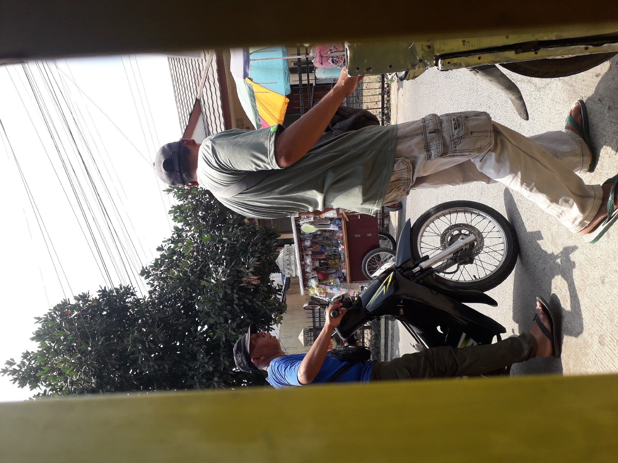 The image captures a street scene in Cankuang, Indonesia. In the foreground, a man wearing a blue t-shirt and dark pants is seated on a black motorcycle, looking to his right. He is wearing a black cap and flip-flops. To his right, another man, also wearing a cap, is standing and walking alongside the motorcycle, looking forward. This second man is dressed in a light green t-shirt and light-colored cargo pants, and he is wearing flip-flops. He has a dark bag slung across his body. Behind the motorcycle, a food cart is visible, displaying various items for sale. The cart has colorful umbrellas providing shade. In the background, there are trees, a tiled roof house, and utility lines overhead. The scene appears to be taking place on a paved road with shadows cast on the ground.
