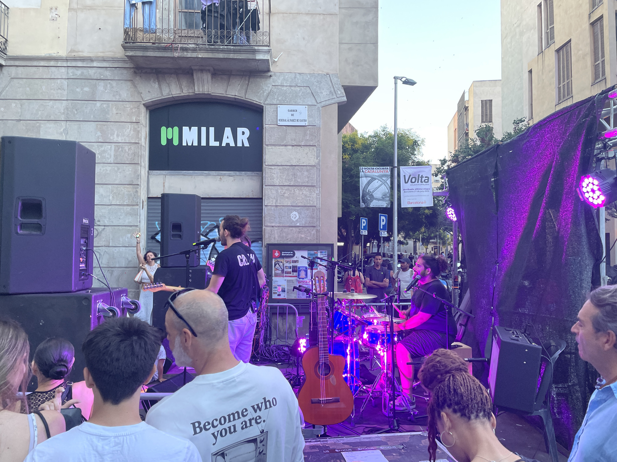 The image captures an outdoor music performance in Barcelona, Spain, likely in a street or plaza setting. In the foreground, several people are partially visible, facing away from the camera and towards a stage. On the stage, a band is performing. A male drummer is seated at a drum kit, illuminated by purple stage lights, and appears to be singing into a microphone. To the left of the drummer, a male guitarist is standing, holding an electric guitar and also singing. A female singer in a white dress is visible in the background, to the left of the stage, with her arm raised. Behind the stage, a black tarp with purple lights creates a backdrop.
The urban environment is evident with buildings visible behind the stage. A sign on a building reads "MMILAR" in green lettering. Another sign above an arched doorway indicates "Carrer de General Alvarez de Castro," confirming the location as Carrer de General Álvarez de Castro in Barcelona. Streetlights and trees are also present in the scene. The overall atmosphere suggests a lively public event.