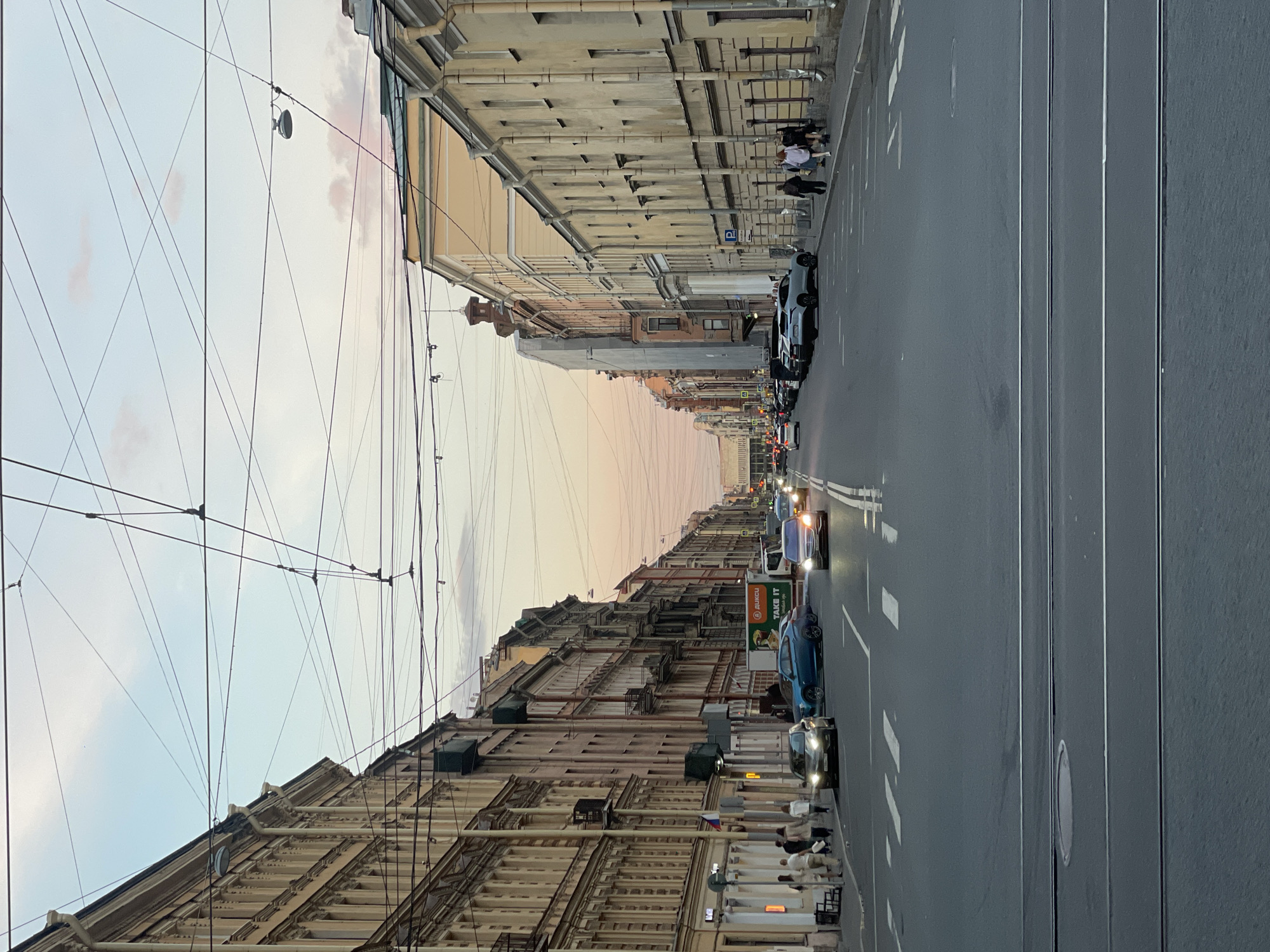 The image displays a wide urban street in Saint Petersburg, Russia, framed by multi-story historical buildings under a twilight sky. The perspective looks down the long thoroughfare as it recedes into the distance. The sky above transitions from a light blue at the zenith to soft pink and orange hues near the horizon, indicating either dusk or dawn. A dense network of overhead electrical wires crisscrosses the sky, characteristic of cities with tram or trolleybus systems.
On both sides of the street, tall buildings with intricate facades, cornices, and numerous windows line the roadway. A small Russian flag is visible on the facade of a building on the left side. The asphalt road features multiple lanes with clear white markings, and tram tracks run along the right side.
Several vehicles are present on the street; some cars are in motion with their headlights on, while others are parked along the sidewalks. Notable vehicles include a light blue car and a black car in the mid-ground. People are visible on both sidewalks, with a group gathered on the right side near parked cars. A blue parking sign with a white 'P' symbol is also seen on the right sidewalk. A billboard on the left side of the street displays partial text "2ГИС" and "TAKE IT". The scene captures everyday city life during the transitional hours of the day.