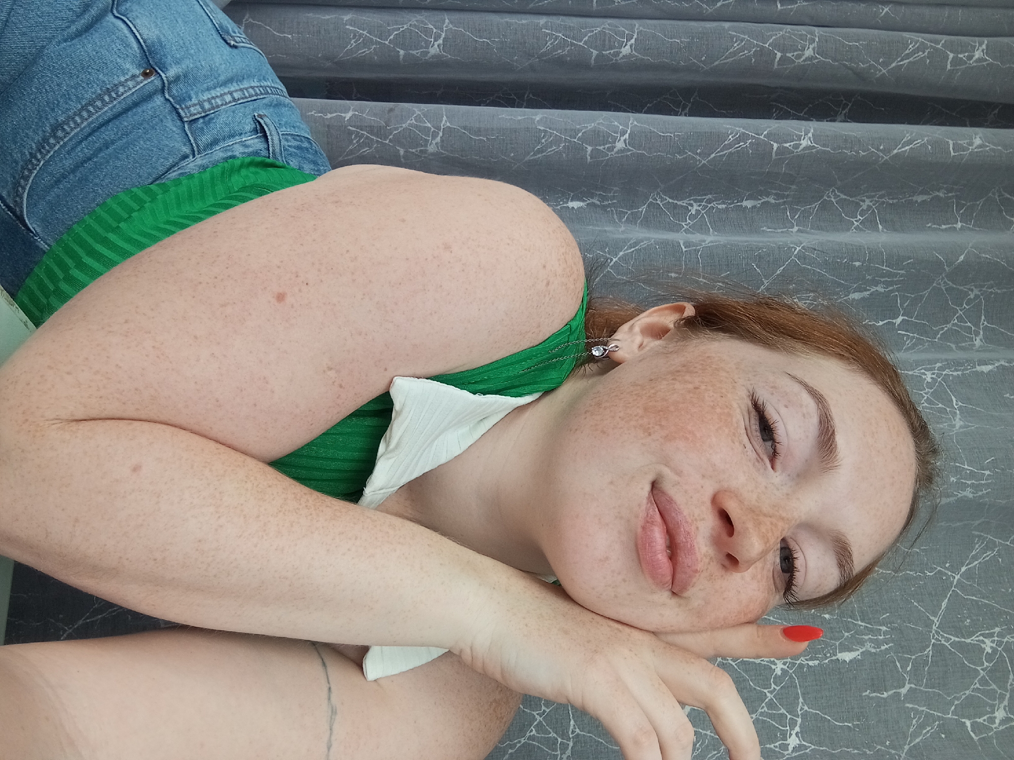 A fair-skinned, red-haired woman with prominent freckles lies on a gray, patterned surface. She is wearing blue denim shorts and a green and white top. She is wearing a dangling earring. The location is not discernible from the image and cannot be identified as Yurgamysh, Russia.
