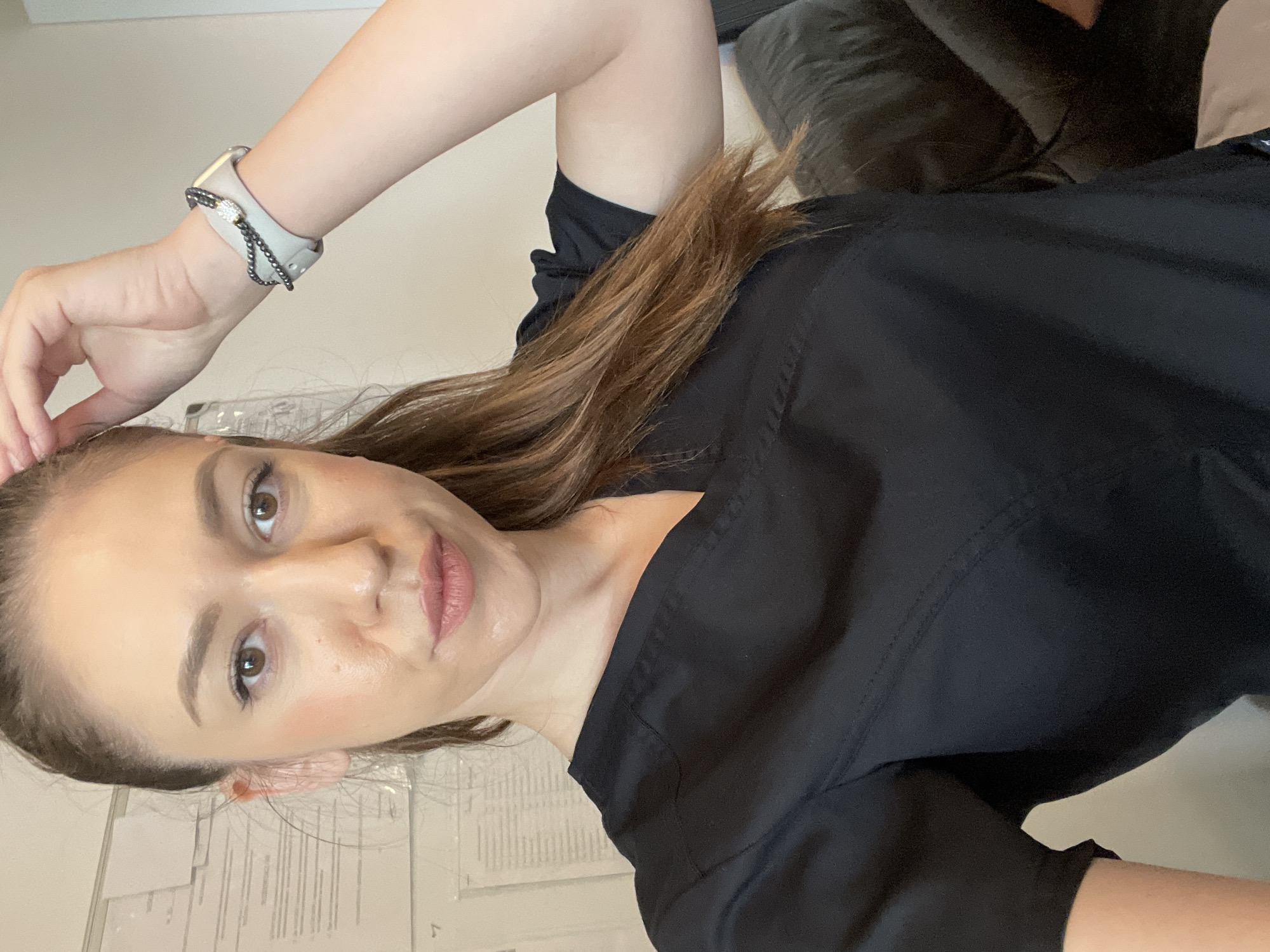 A woman with light brown hair is posing for a selfie. She is wearing a black uniform and a silver smartwatch with a decorative band. She is indoors, with a glimpse of a grey couch in the background and papers on the wall. There is no information about the city of Sofia, Bulgaria.
