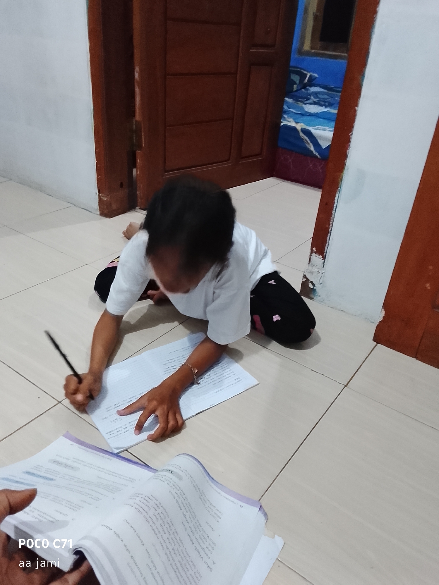 A young girl is kneeling on a tiled floor, writing on a piece of paper with a pencil. She is wearing a white t-shirt and black pants with pink accents. She has dark hair and is focused on her work. In front of her is another open book. The room appears to be a bedroom, with a bed visible in the background, covered with a blue and white patterned comforter. A wooden door is also prominent in the scene. The flooring is light-colored tile. The scene is indoors, likely in a residential setting in Tasikmalaya, Indonesia.