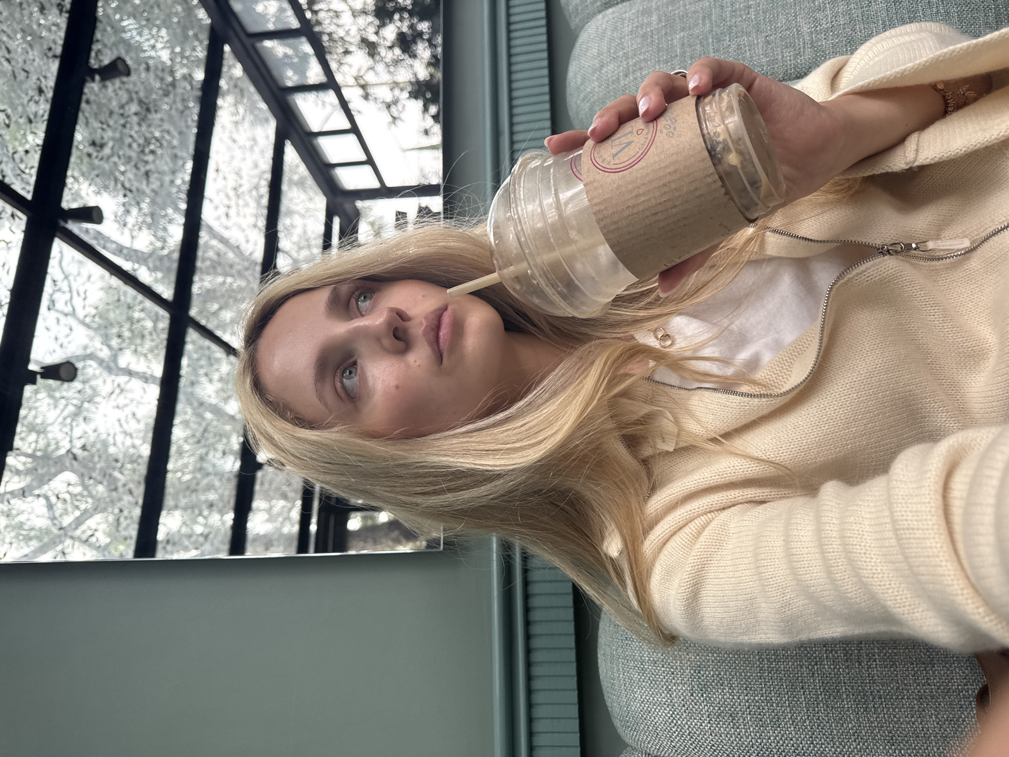 A young woman with blonde hair sits on a teal couch and holds a plastic cup with a straw. She is wearing a cream-colored zippered cardigan over a white t-shirt. The background appears to be a modern cafe with a glass ceiling showing tree branches and a mirror reflecting the interior. The overall atmosphere is casual and relaxed. The location is in Mexico City, Mexico.