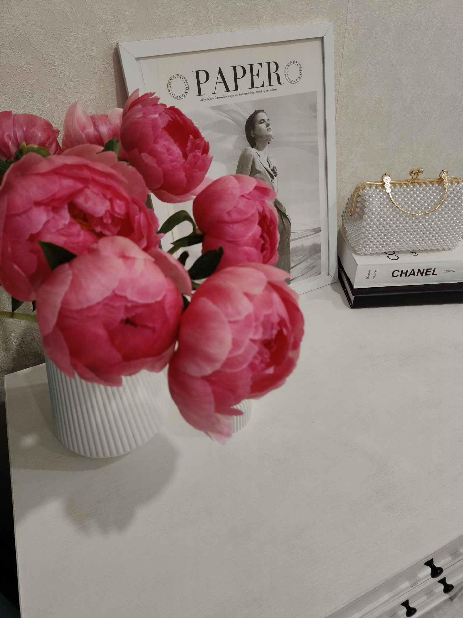 The image depicts an indoor still life arrangement on a light-colored table against a textured, light beige wall in Unknown, Unknown. In the foreground on the left, a white ribbed vase holds a bouquet of approximately six to seven vibrant fuchsia-pink peonies, some partially open and some fully bloomed, with dark green leaves interspersed. A prominent shadow of the vase and flowers is cast on the table surface. Towards the right, a small stack of two books is visible; the top book is white with "CHANEL" printed in black, and the bottom book is black. Resting on the books is a small, white clutch bag completely covered in pearls, featuring a gold-colored frame, clasp, and small handle. In the background, behind the flowers and books, a white-framed picture or poster is displayed against the wall. The poster's visible text includes "PAPER" in large bold letters at the top, and below it, "All products featured on Vogue are independently selected by our editors." The image within the frame shows a monochrome photograph of a woman in a suit, looking upwards. The wall has a subtle, light-colored textured pattern. No people are present in the scene.