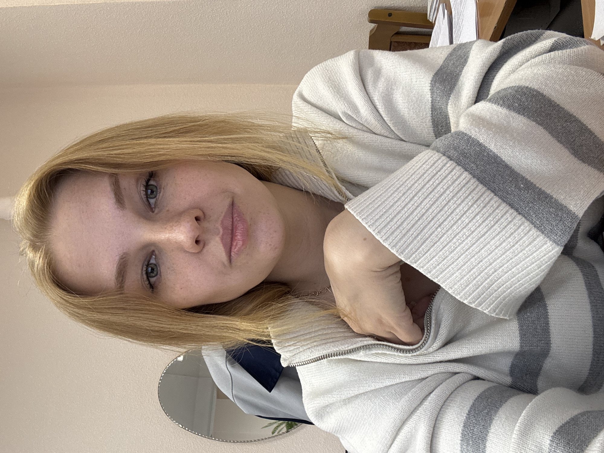 A fair-skinned young woman with blonde hair is facing the camera. She has light eyes and freckles, and her lips are closed in a neutral expression. She is wearing a white striped sweater with grey stripes and a zipper. The sweater is partially zipped. Her hand is visible near the zipper. The background is a plain wall, and there is a round mirror visible behind her left shoulder. A partial view of furniture is visible in the upper right corner. The location is not discernible from the image.