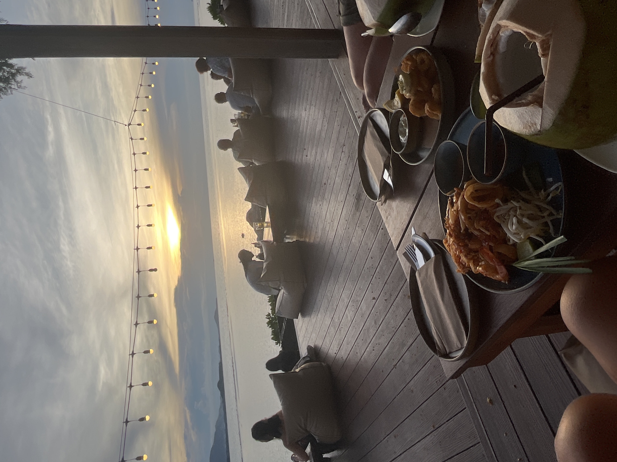 The image depicts a serene outdoor dining scene at sunset on a wooden deck overlooking the ocean in Baan Plai Laem, Thailand. A string of incandescent lights is strung across the sky, illuminating the tranquil water and distant islands. Several people are seated on large, cushioned floor seats, enjoying the view. In the foreground, a table is set with plates of food, including what appears to be pad thai and fried calamari, accompanied by dipping sauces and a halved coconut with a straw. The overall atmosphere is one of relaxation and enjoyment of a beautiful evening meal.
