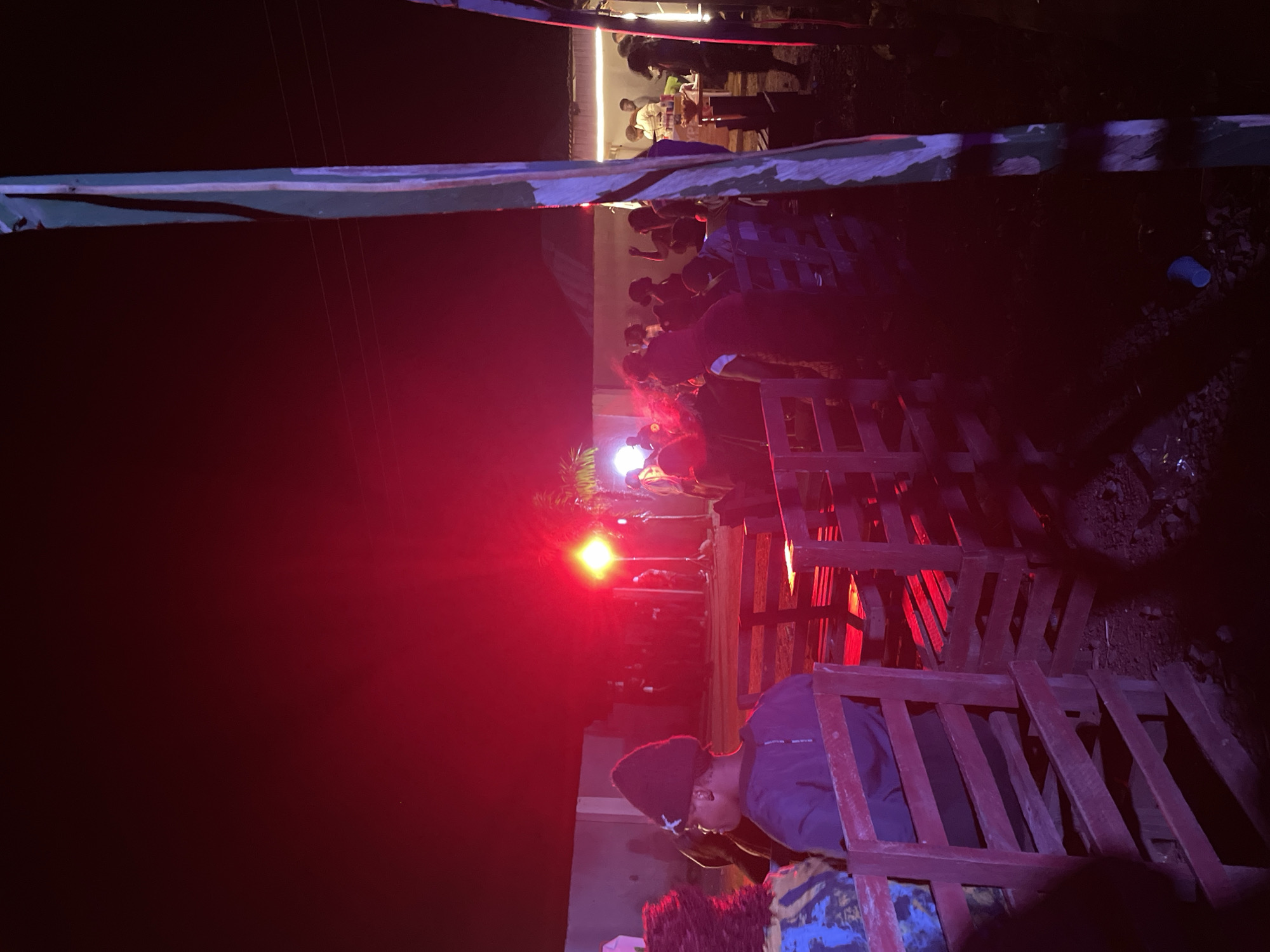 The image depicts an outdoor gathering at night, likely a social event or party, taking place in Durumi, Nigeria. Intense red lighting dominates the scene, casting a crimson glow on the surroundings and illuminating a crowd of people. In the foreground, wooden benches, possibly repurposed pallets, are arranged in rows, suggesting a seating area. A person wearing a dark beanie and a dark t-shirt is seated, facing away from the camera, looking towards the main gathering. The background reveals a wall and structures, typical of a residential or commercial area in Durumi. Several individuals are visible in the distance, some appearing to be standing and socializing, while others are seated. A bright, intense light source, likely a spotlight or stage lighting, is positioned in the middle ground, contributing to the dramatic red illumination. The ground appears to be unpaved, with some debris visible. A tall, vertical structure, possibly a pole or part of a temporary setup, with alternating green and white stripes, stands prominently on the right side of the frame. The overall atmosphere suggests a lively and energetic event.