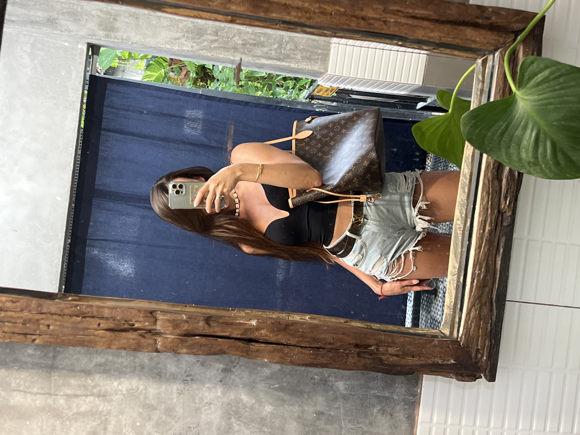 In a bathroom with a rustic, natural aesthetic, a woman stands in front of a large, wooden-framed mirror, taking a selfie. The mirror's frame is made of weathered, natural wood, adding a raw, organic element to the scene. The woman is wearing a black crop top, light-wash ripped denim shorts, and a designer belt. She is holding a brown designer tote bag. The background visible in the mirror includes a dark blue curtain, greenery, and white tiled walls. The overall impression is one of casual style and a relaxed atmosphere, possibly in a boutique hotel or a trendy restroom.