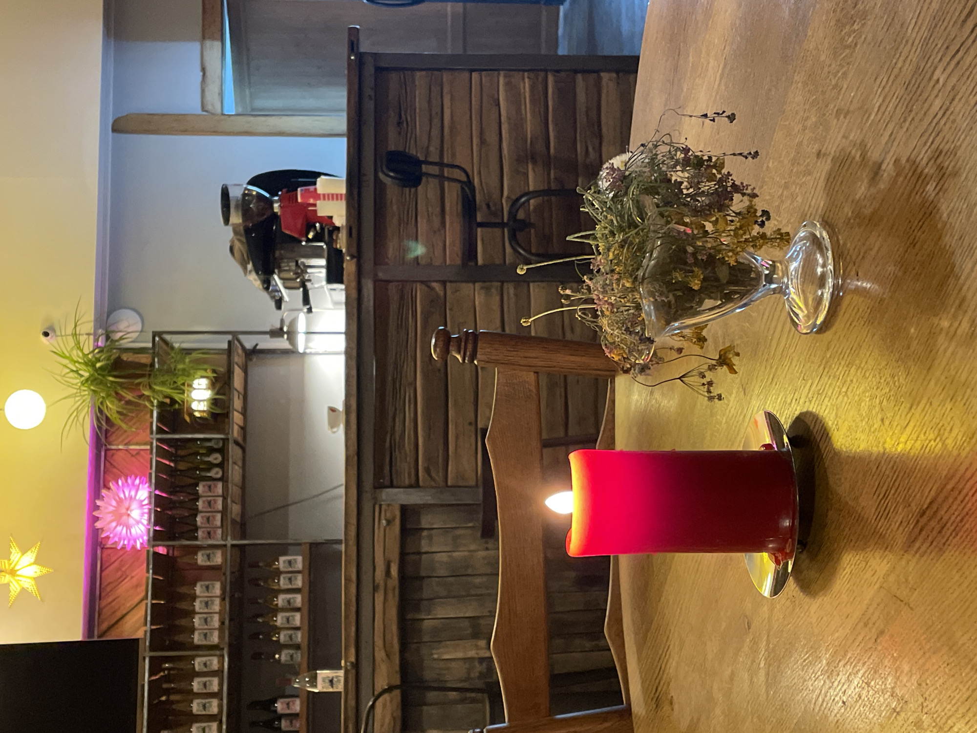 The image depicts an interior scene, likely a cafe or restaurant, with a focus on a wooden table in the foreground. A large, red, lit candle sits on the table, casting a warm glow and a distinct shadow. To the right of the candle, a glass vase holds a bouquet of dried flowers and plants. In the background, a wooden bar with shelves stocked with bottles is visible. A coffee machine is situated behind the bar, and a bar stool is placed in front of it. The lighting is dim and atmospheric, with overhead decorative lights adding to the ambiance. The overall impression is one of a cozy and intimate setting. The location is specified as Stepantsminda, Georgia.