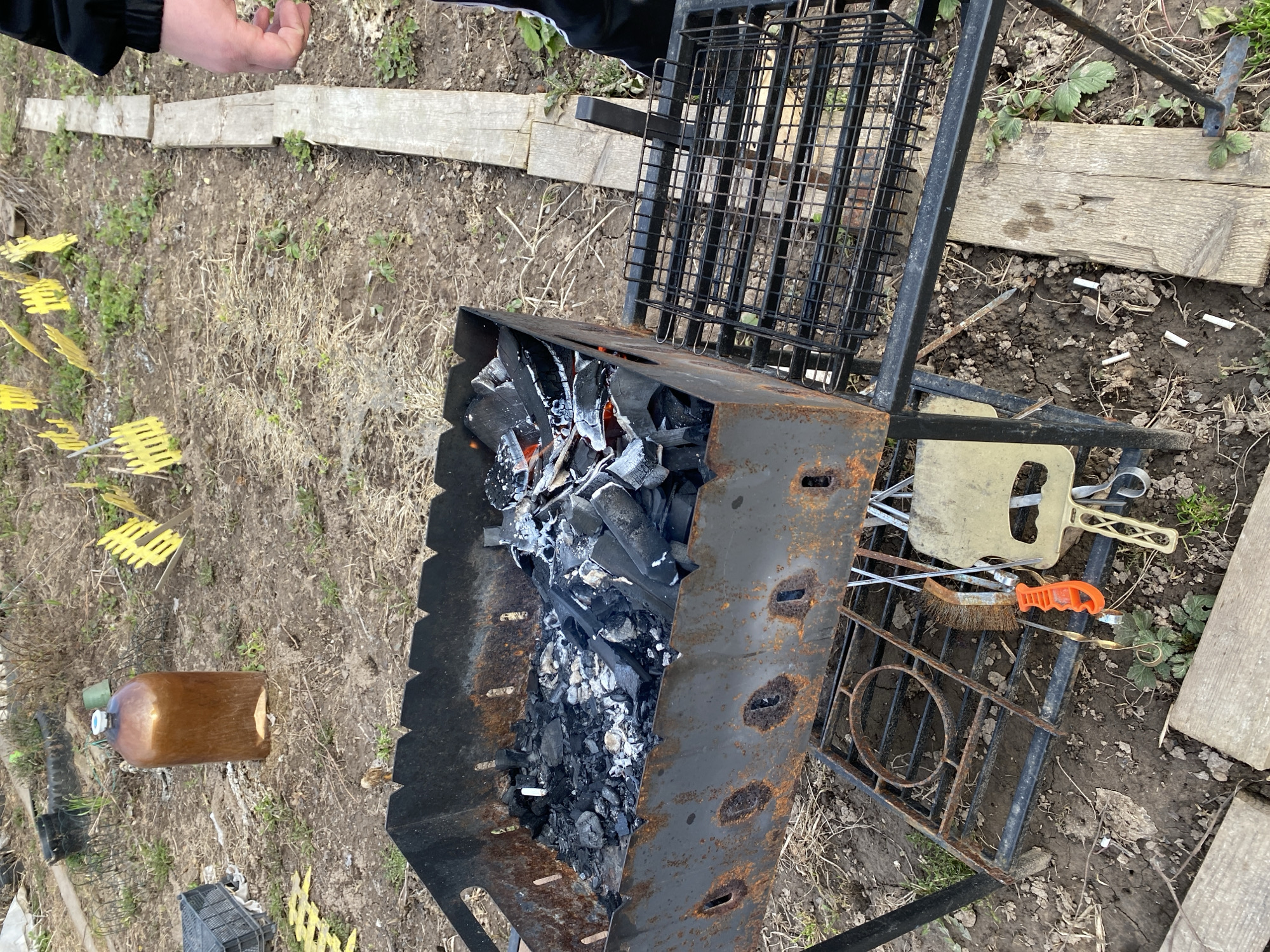 An overhead view captures an outdoor scene, likely a backyard or rural setting in Фоки, Russia, centered on a lit barbecue setup. A rusty, rectangular metal brazier holds glowing embers and black charcoal, indicating an active or recently started grilling session. A black metal grill grate rests on top of the brazier. To the right, another black metal stand or grate with a circular design lies on the ground, serving as a holder for various barbecue tools including a light-colored spatula, a brush with an orange handle, and metal tongs.
The ground is a mix of bare dirt, patches of dry grass, and some green vegetation, appearing somewhat natural or unkempt. Several wooden planks are laid irregularly across the ground. In the left foreground, multiple yellow plastic garden markers or labels are scattered, though their text is unreadable. A brown plastic bottle with a light green cap lies horizontally near these markers. A person's hand and forearm, wearing a black sleeve, are visible in the upper left corner, suggesting human presence and activity related to the barbecue. Several white cigarette butts are scattered on the ground near the brazier. The natural light indicates it is daytime, without strong shadows to suggest a specific time or intense sunlight.