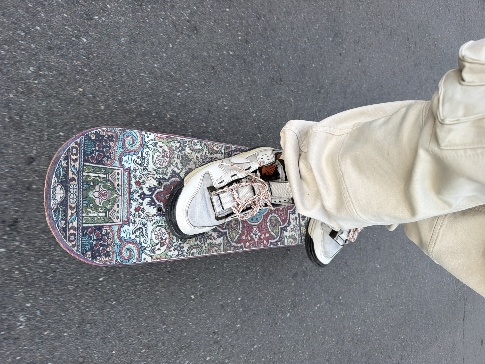 A skateboard with a patterned deck sits on asphalt, with a person standing on it. The person wears light-colored cargo pants and white sneakers with patterned laces. The background is blurred, suggesting motion. No evidence indicates the location is Moscow, Russia.