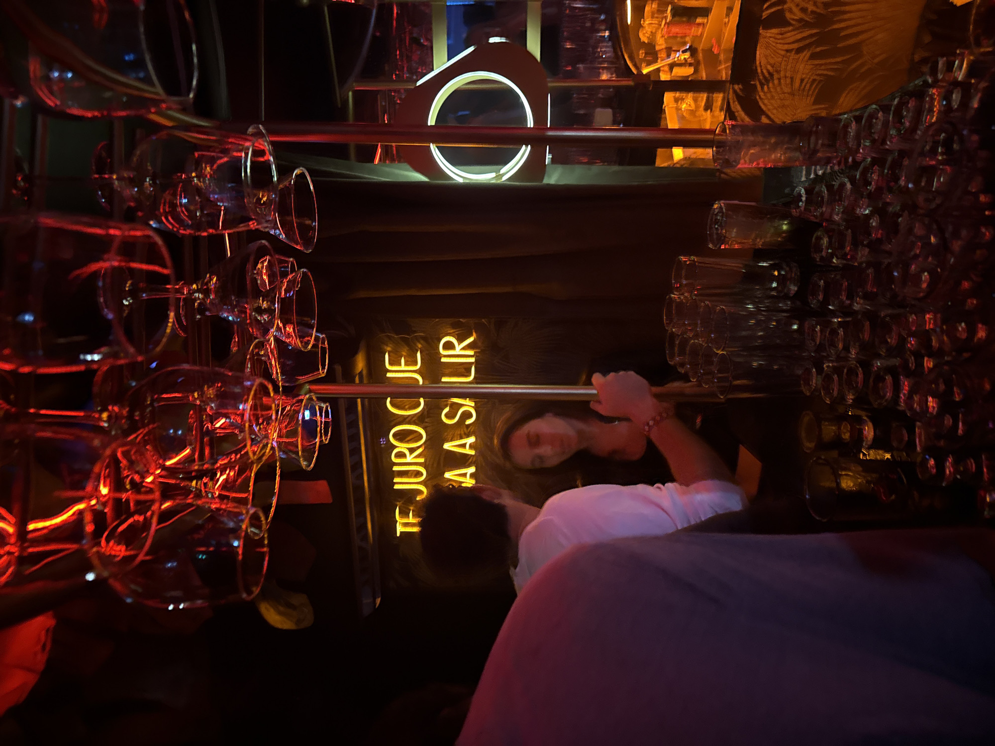 The photograph, taken indoors, shows a dimly lit bar setting in Mexico City, Mexico. The viewpoint is positioned behind two individuals at the bar. One individual wears a white shirt, with only the back of their head and shoulder visible. To their left, a woman is seen holding onto the bar, wearing a black shirt and a red bracelet. A glowing yellow neon sign is visible in the background, with text that appears to read "TE JURO QUE VUELVA A SALIR." The bar is adorned with rows of wine glasses suspended upside down from racks, illuminated by a red light. Further back, there are rows of clear glasses and bottles. There is a circular lamp emitting a white light positioned higher up in the frame. The overall atmosphere suggests a lively, intimate bar environment.