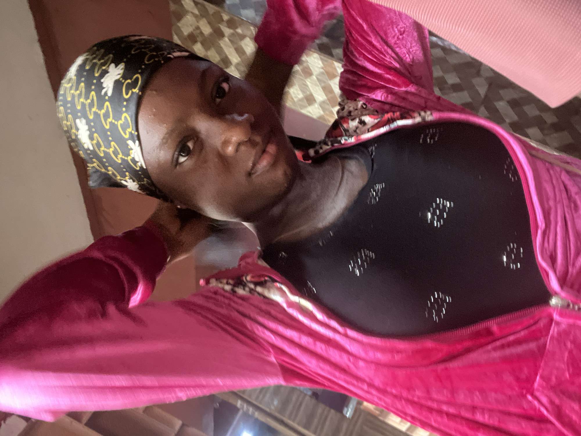 In an indoor setting, a person with dark skin and wearing a pink velour tracksuit over a black top with a patterned headscarf poses for a selfie. The person's hands are raised to the back of their head, and their gaze is directed at the camera. The background features a wall with a patterned texture and what appears to be a wooden partition, suggesting a domestic environment. The lighting is ambient and originates from the front, illuminating the subject. The location is identified as Nkpor, Nigeria.