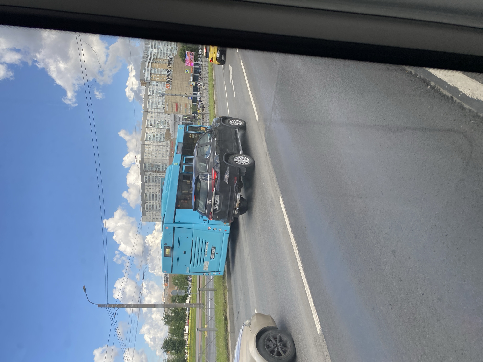 The image shows a bright, sunny day in Saint Petersburg, Russia, with a wide road and blue sky filled with white clouds. A bright blue public transport bus is visible in the middle of the frame, with its rear facing the camera. Next to it, a black SUV is driving in the same direction. Further down the road, a yellow car is also visible. The scene appears to be in a populated area, with tall apartment buildings in the background, featuring a variety of architectural styles and colors. Some trees and streetlights are also present. The overall atmosphere is one of urban activity on a pleasant day.