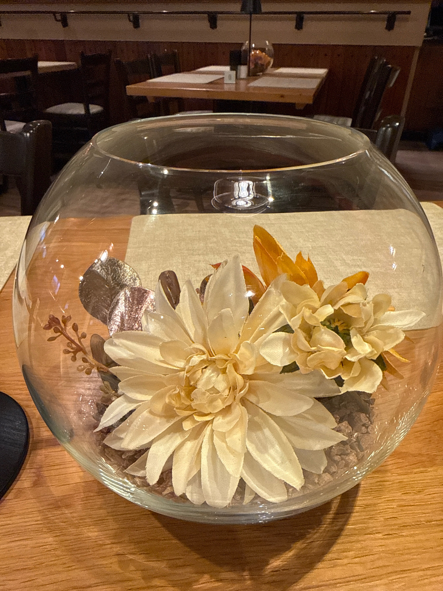 The image shows a close-up view of a glass bowl containing artificial flowers and decorative elements, placed on a wooden table. The bowl is filled with small brown pebbles, and a large, pale yellow dahlia-like flower is prominently featured, along with a smaller, similar flower and some autumnal-colored leaves and twigs. The background is blurred, suggesting an indoor setting like a restaurant or dining area, with tables and chairs visible. Reflections on the glass indicate artificial lighting. There are no people visible in the image. The specific location in Bonn, Germany cannot be determined from the visual information provided.
