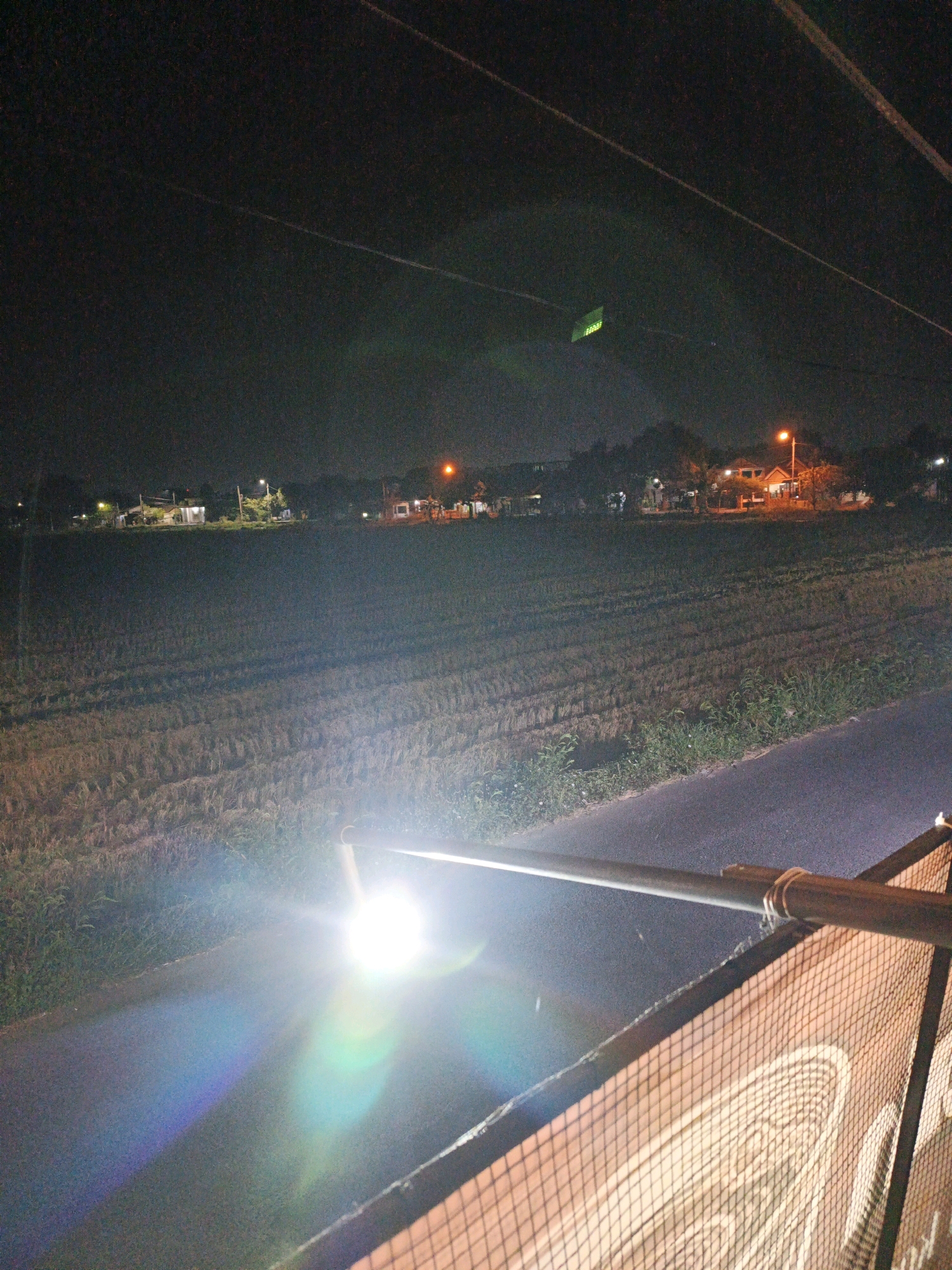 The image depicts a nighttime scene in Ketegan, Indonesia. The foreground shows a bright, lens-flared light source illuminating a road and a fenced structure. The road appears to be asphalt, and the fence is made of metal mesh. In the middle ground, a harvested agricultural field is visible, with rows of stubble indicating where crops were previously grown. Beyond the field, a cluster of houses with illuminated windows and streetlights line the edge of a treeline. The sky is dark, with visible power lines stretching across the frame. A small, green, rectangular object is visible in the upper portion of the sky, possibly a drone or kite.