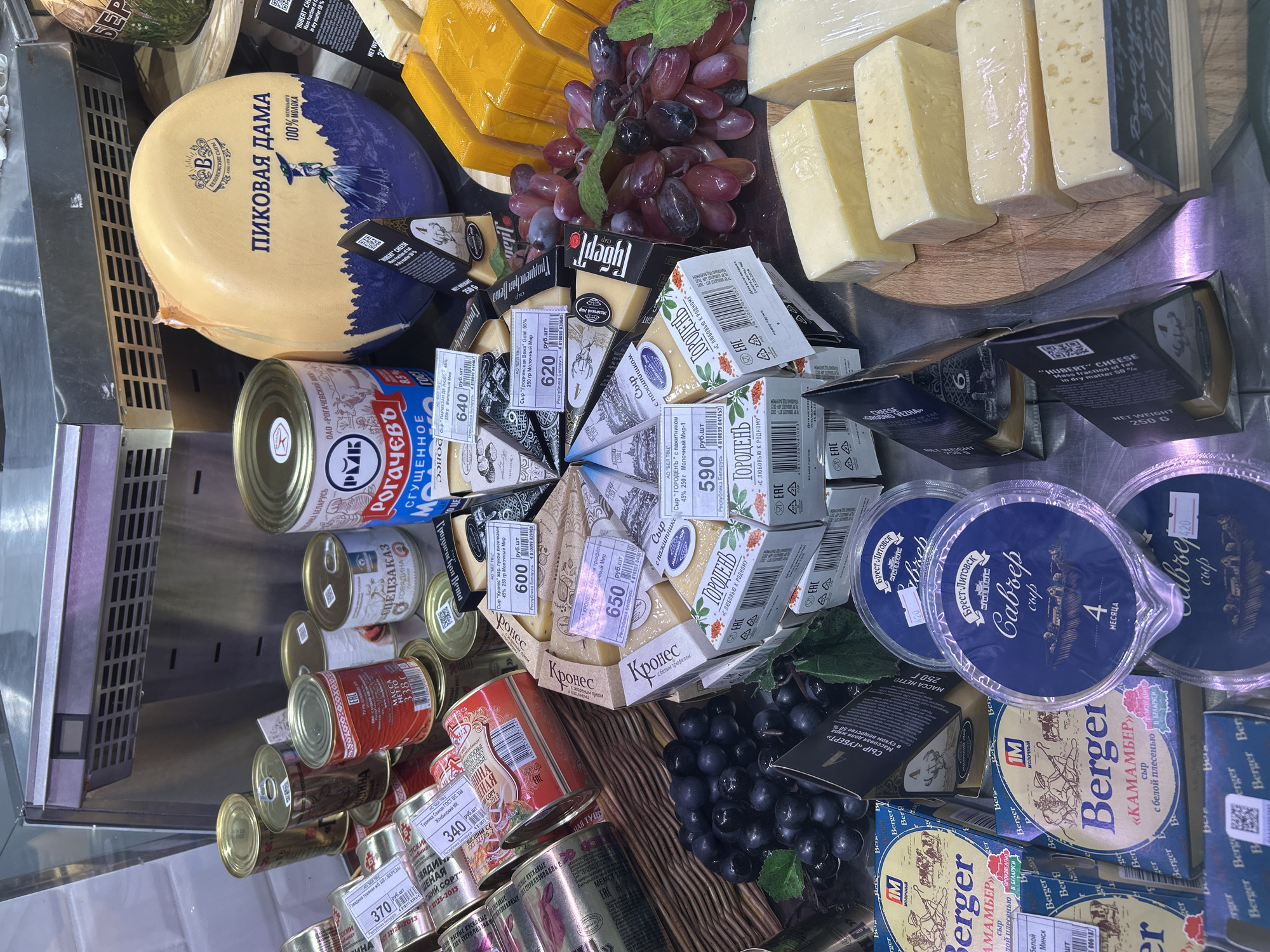This is an eye-level close-up shot of a display case filled with various food items, likely in Moscow, Russia based on the product labeling. There are several types of cheese including a large wheel of "Pikovaya Dama", triangle-shaped cheese wedges in packaging labeled "Гродненсь", and packaged Camembert cheese labeled "Berger". Grapes (both red and dark blue/black) are present, likely as a cheese accompaniment.
Canned goods are stacked on the left side of the display, including canned meat ("Говядина тушеная") and condensed milk ("Рогачевъ"). The prices are displayed on the cheese packaging, indicating they are for sale. The overall environment suggests a grocery store or deli counter. The scene is static, showing a selection of food products arranged for sale.