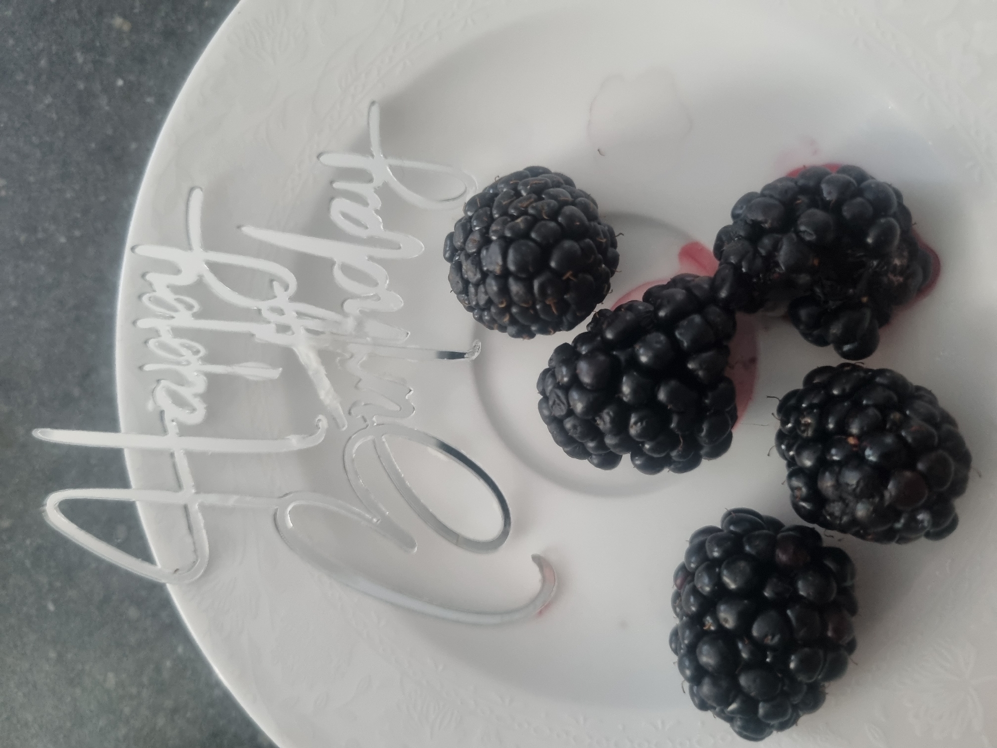 A close-up, high-angle shot shows a white plate on a textured, dark gray surface, possibly a countertop in Samara, Russia. On the plate, five ripe blackberries are arranged in a cluster, with some pink juice seeping from two of them. A silver cake topper, spelling out "Happy Birthday" in cursive, is positioned behind and above the blackberries, partially obscuring them. The lettering is thin and reflective. The lighting appears to be natural, coming from above and to the left, casting subtle shadows.