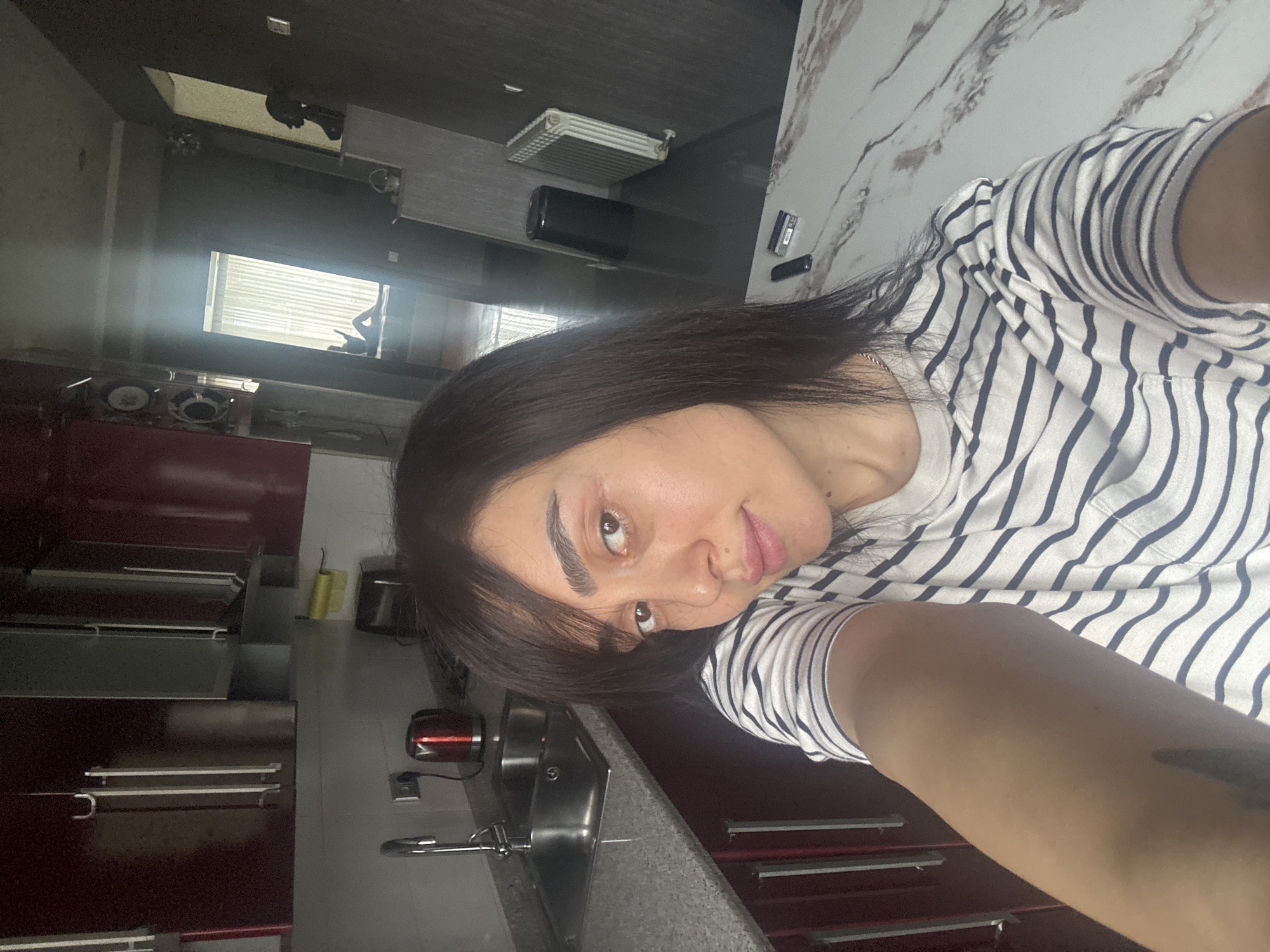 A person is taking a selfie in what appears to be a kitchen. The person has dark hair and is wearing a striped shirt. Visible in the background are kitchen cabinets, a sink, and a window. Based on the style of the kitchen and available light, the location is most likely indoors. There is no discernible information in this image to place the setting in Tbilisi, Georgia.