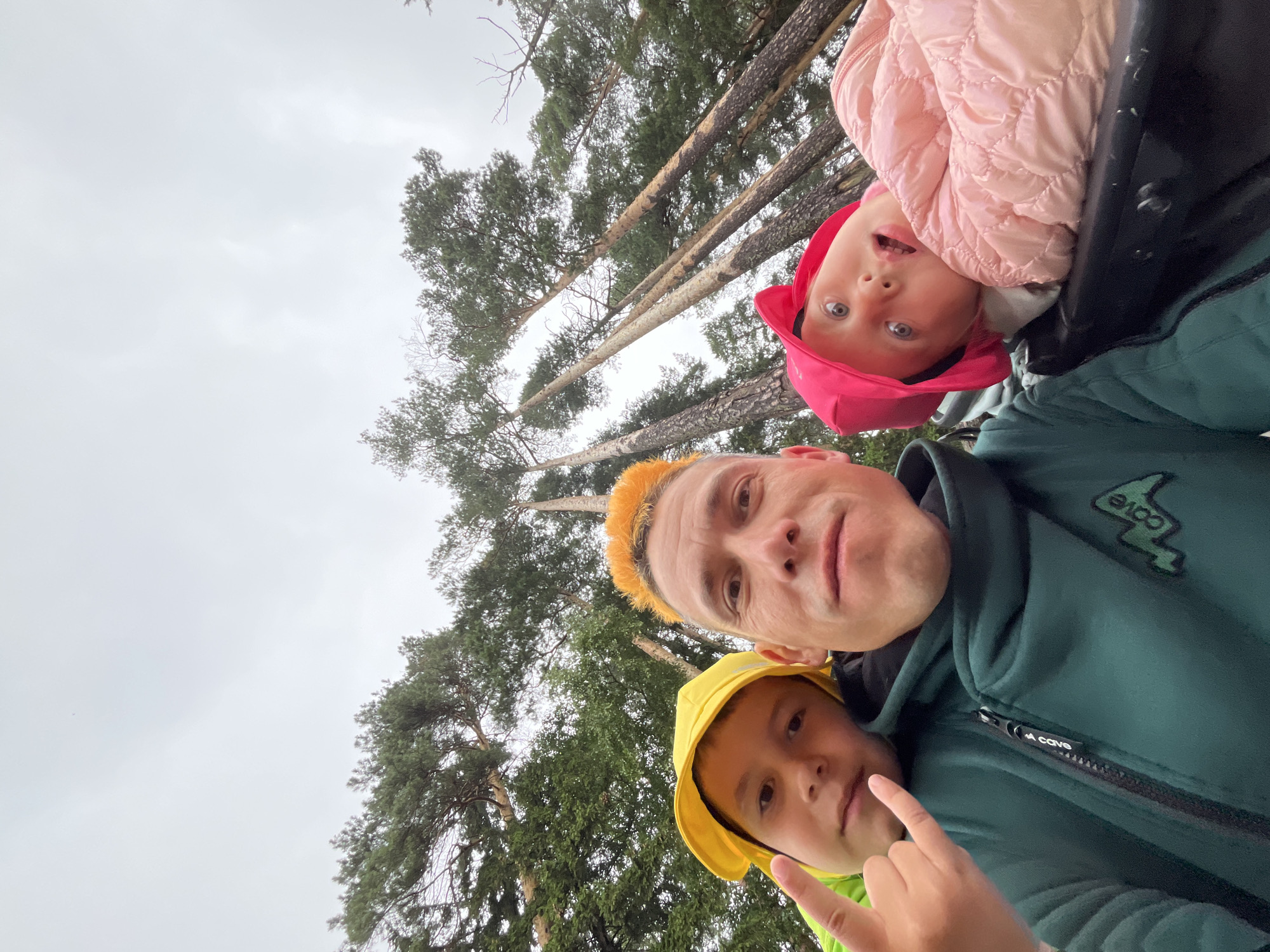In a selfie taken outdoors with a background of tall pine trees and an overcast sky, three individuals are visible. The person in the foreground, in the center, has bright orange hair and is wearing a dark green hooded jacket with a zippered front. To their left, a young boy wearing a yellow hat and a bright green jacket is giving a rock-and-roll hand gesture. To the right of the central figure, a young child wearing a pink hat and a pink quilted jacket looks towards the camera with an open mouth. The overall scene appears to be in a wooded area, possibly a park or forest, with the towering trees suggesting a natural setting. There are no discernible landmarks or specific urban features that would indicate a particular location within Chaikovsky, Russia.