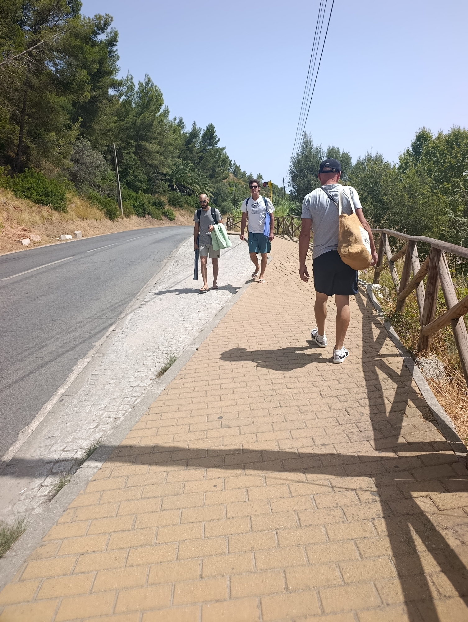 Three men walk along a paved path next to a road lined with trees and vegetation. The man in the foreground, facing away from the camera, wears a grey t-shirt, dark shorts, and a baseball cap, carrying a beige tote bag over his shoulder. He is followed by a man in a white t-shirt and blue shorts, carrying a blue bag. Further ahead, a third man in a grey t-shirt and plaid shorts walks with a green towel over his arm and a dark object, possibly a rolled-up mat, in his hand. The path is constructed of yellow bricks, and a wooden fence runs along the right side of the path. The road to the left is asphalt, with a stone border. The sky is clear and blue, and the sun casts long shadows. The presence of pine trees and the arid vegetation suggests a Mediterranean climate. The scene appears to be a pathway leading to or from a beach or recreational area.