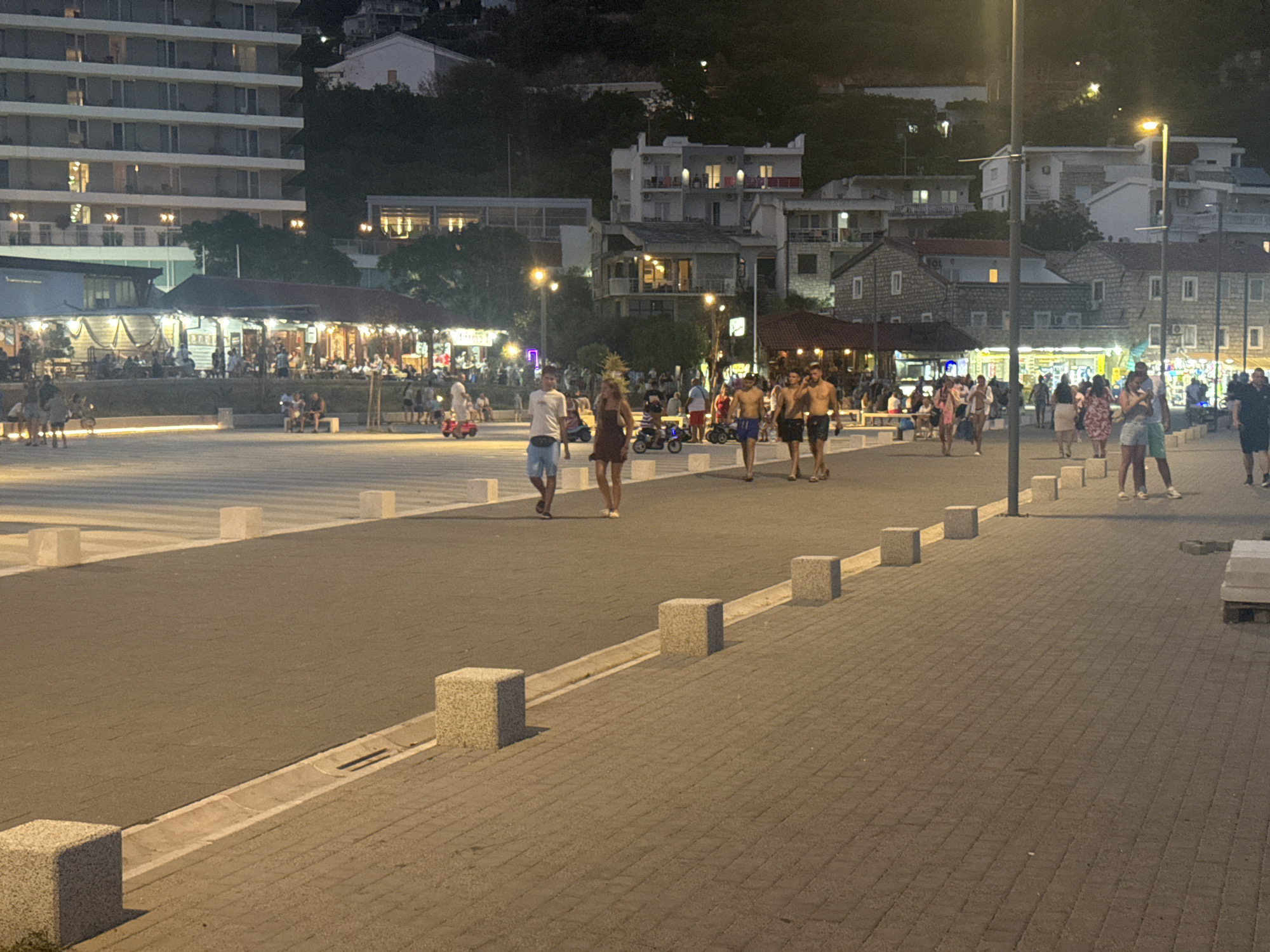 The image depicts an evening scene in Sutomore, Montenegro. A wide paved promenade is visible in the foreground, lined with stone cube bollards. Several people are walking on the promenade. In the background, a row of restaurants and shops with lit facades line a street. Behind the buildings, a hillside is visible with more structures and trees, illuminated by artificial lights. The overall atmosphere is that of a busy tourist area at night.