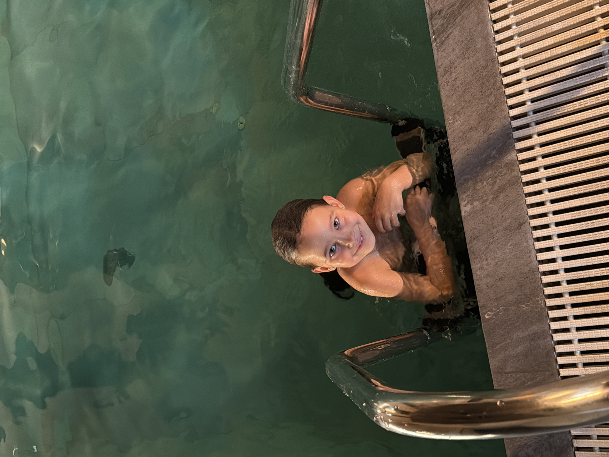 A fair-skinned child with short brown hair is in a body of water, likely a pool, with a greenish hue. The child is holding onto a metal railing along the edge of the pool, which has a dark gray stone border adjacent to a white slatted surface. The child is looking directly at the camera and smiling. The environment appears to be indoors, and the lighting is dim. There is no information to suggest that the scene is located in Shamoni, Russia.