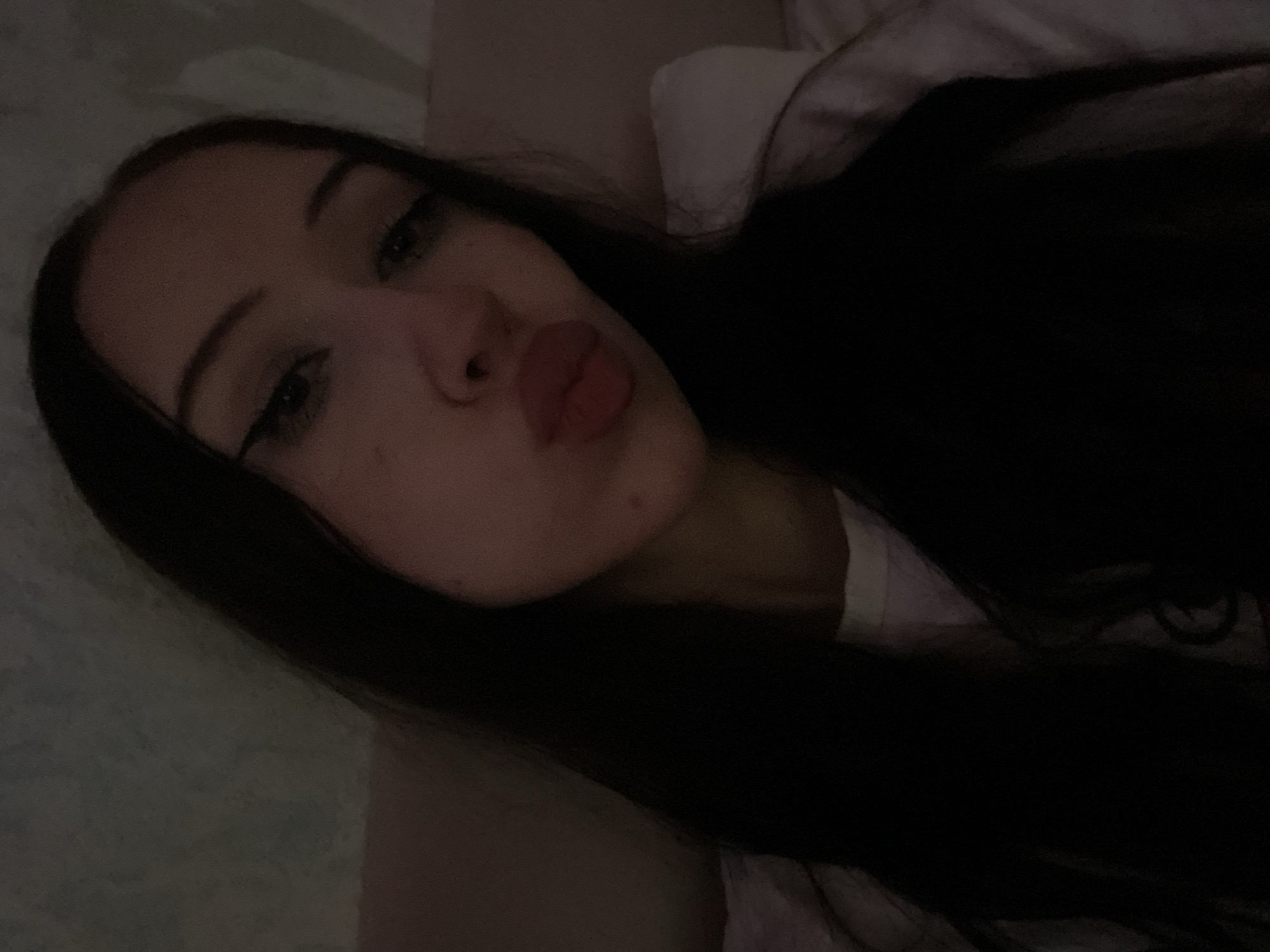 A young woman with long, dark hair and eyeliner is posing for a selfie. She is leaning her head to the left and puckering her lips. The background is dimly lit and appears to be a bedroom with a textured wall and a white pillow. The image is a close-up of her face and upper body. There is no information available to indicate the location in the city of Votkinsk, Russia, or any other specific location.
