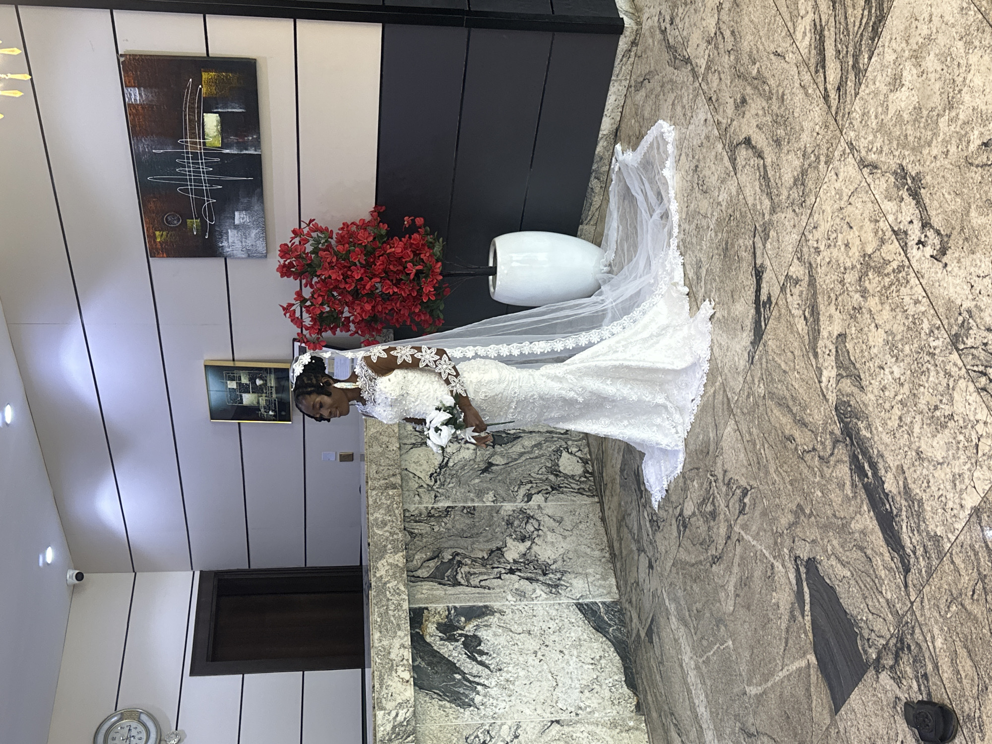 The image captures a full-body shot of a bride in a white, lace wedding dress, standing indoors. She has dark skin and her hair is styled in an updo, adorned with a white veil that extends behind her. She is holding a bouquet of white flowers and looking towards her left. The bride is standing on a marble floor with a patterned design.
Behind her, the setting appears to be a modern interior. To her left, there is a large abstract artwork on a wall with black and white stripes. Next to the artwork, a vase filled with red flowers adds a vibrant splash of color. Further to the left, part of a white ceiling with recessed lighting is visible, along with a security camera. A dark-colored door is also present on the left side of the frame.
The overall impression is that of a wedding day moment, possibly after a ceremony or before a reception, within a well-decorated indoor venue in Abuja, Nigeria. The lighting suggests it's daytime.