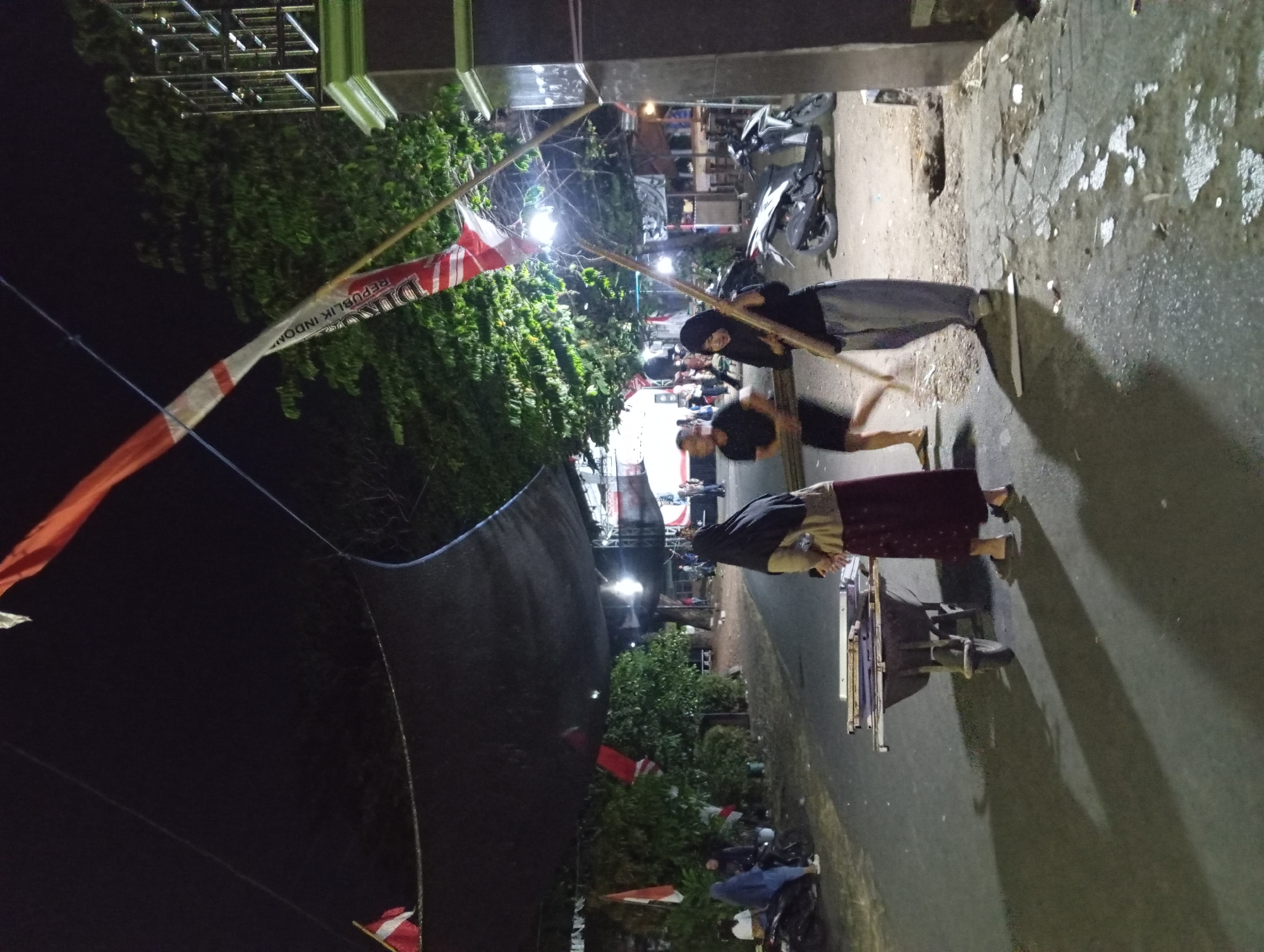 The image depicts an outdoor scene at night in what appears to be a street or communal area. The setting is decorated with Indonesian flags, indicated by the red and white colors and text visible on one of the flags ("REPUBLIK INDONESIA"). Several individuals are present. In the foreground, two women are standing near a wheelbarrow loaded with metallic objects. One woman, on the left, wears a headscarf and a dark red skirt, and is holding a smartphone. The other woman, on the right, wears a black hijab and light grey pants, and is holding a long wooden pole. A man in a black t-shirt and shorts is running in the background, blurred due to motion. Further in the background, there are more people, some seated on motorcycles, and market stalls or temporary structures are visible, suggesting a lively evening event or gathering. A parked motorcycle is also present on the right side of the image. The environment is illuminated by artificial lights, casting long shadows. The presence of flags and what appears to be a gathering point suggests a local celebration or event.
